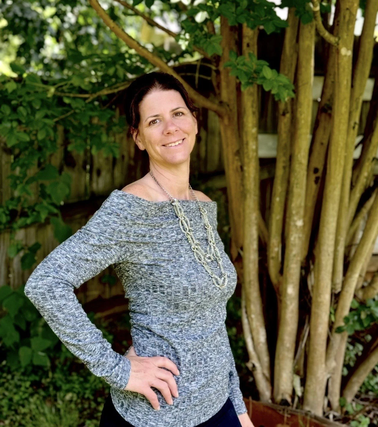 A woman with dark hair and a light gray sweater with a silver necklace, standing outdoors near a tree with green leaves and a wooden fence in the background.