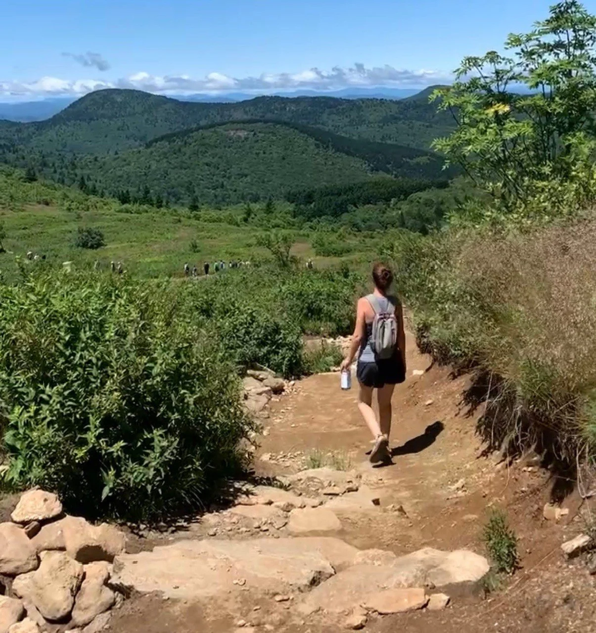 A woman with a backpack walking along a dirt trail in a lush green mountain landscape under a blue sky with clouds.