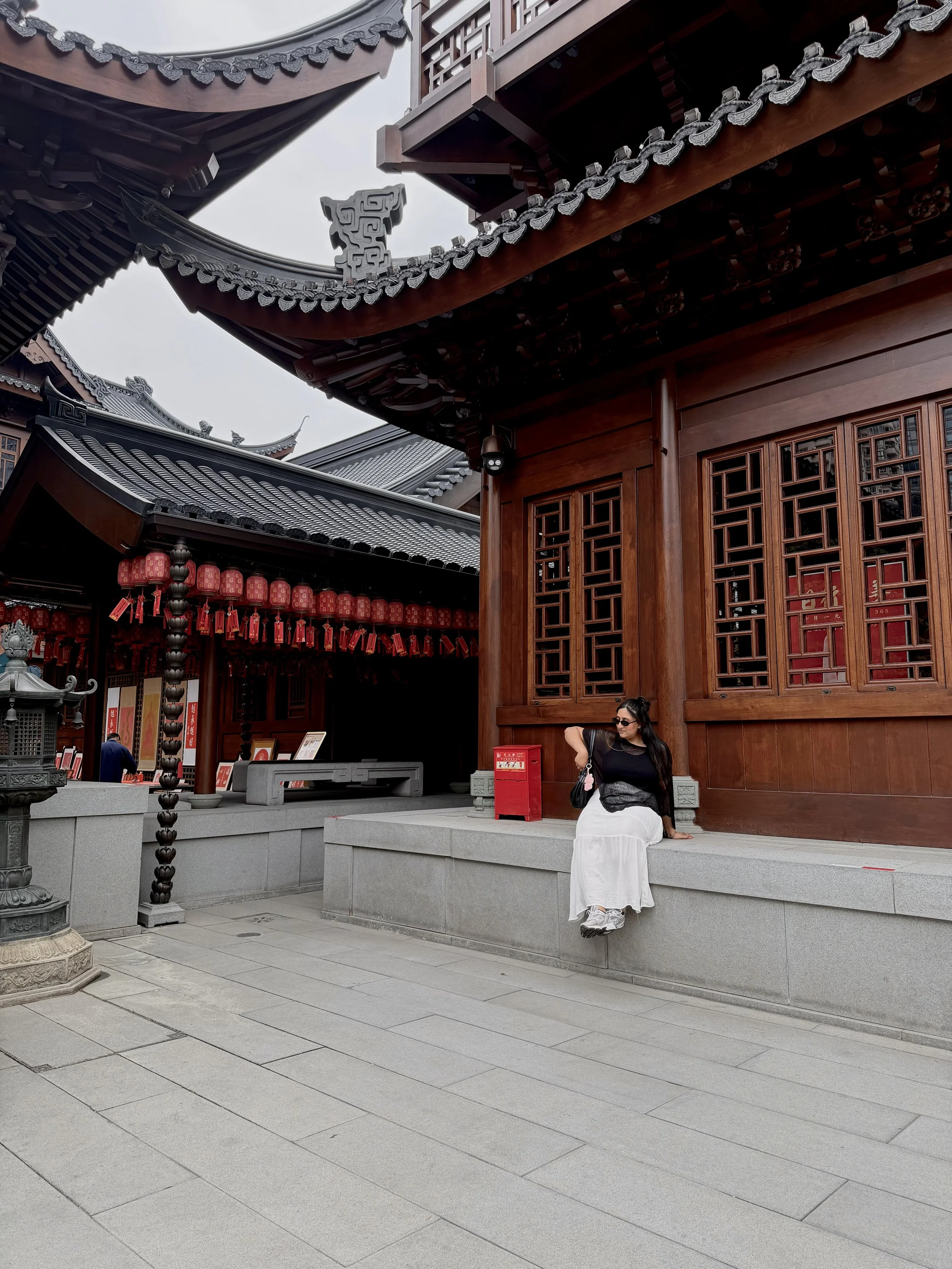 A woman sitting on a stone step outside a traditional East Asian wooden building with ornate roof design and red lanterns.