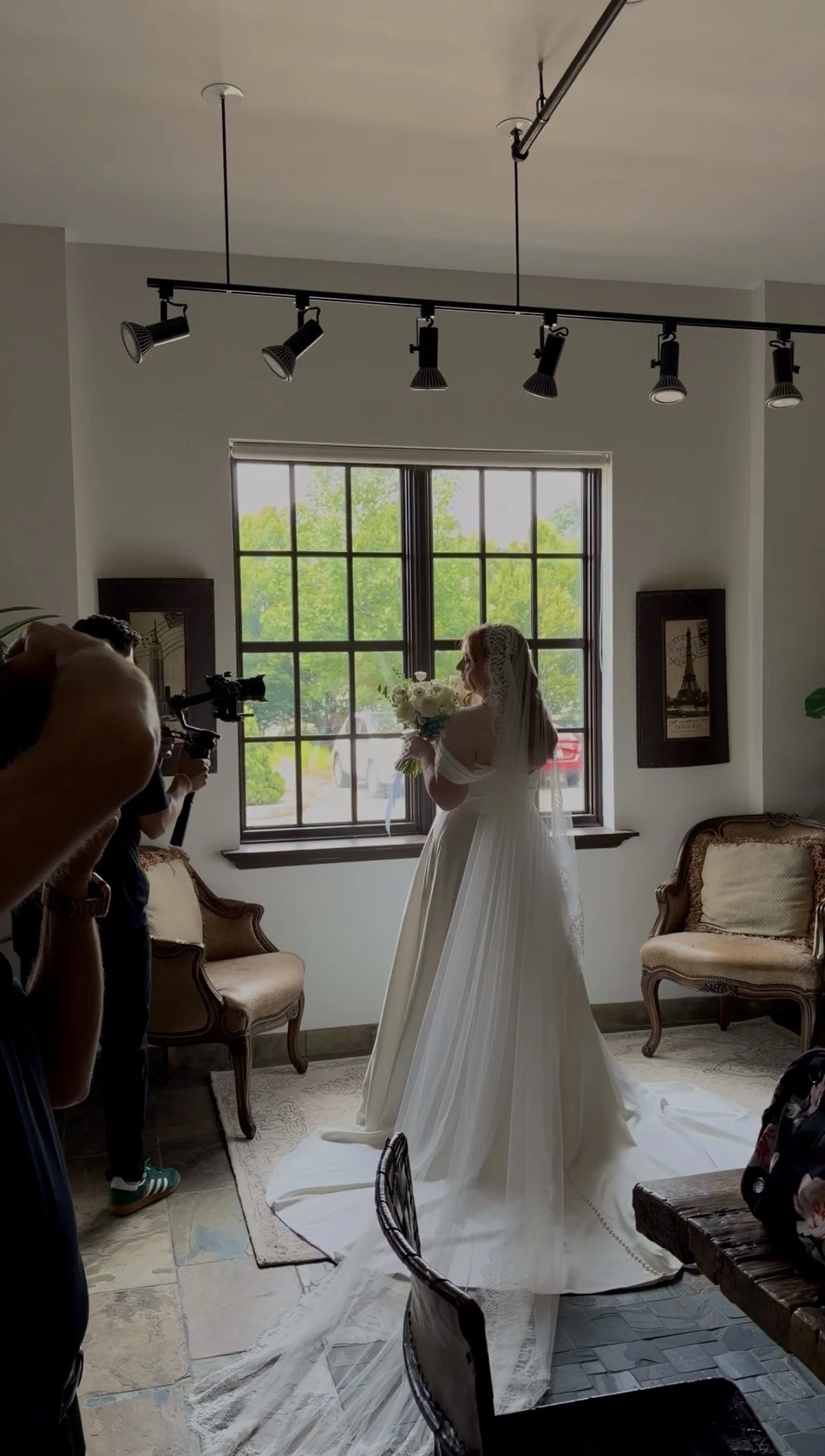 A bride in a white wedding dress holding a bouquet of white roses, standing by a window with green trees outside. Photographers are taking pictures of her in a cozy room with vintage style furniture and framed artwork on the walls.
