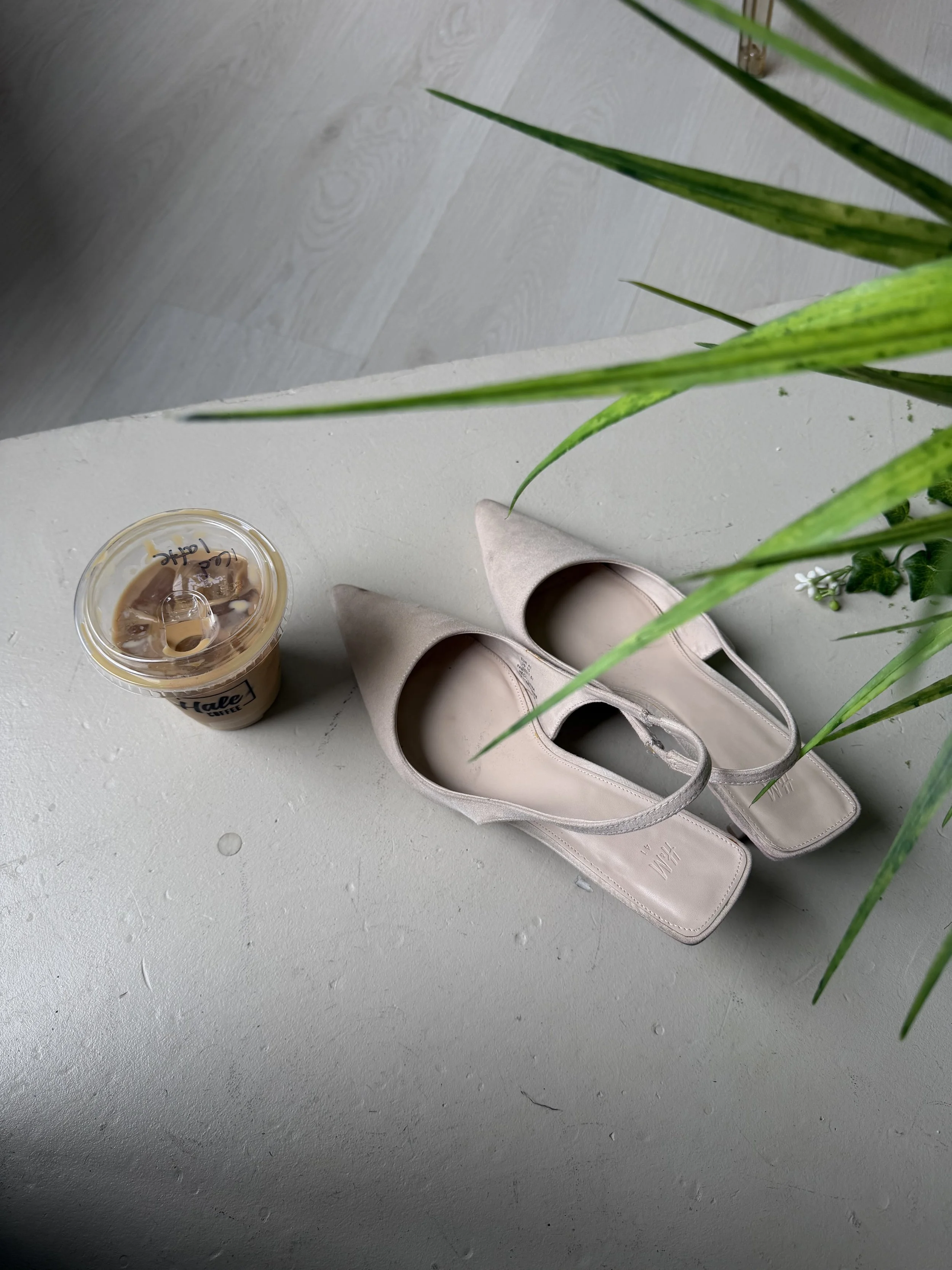 A pair of beige high-heeled shoes, a plastic cup with iced coffee, and a pair of pink flats are on a white textured surface. There is a green plant with long leaves partially visible at the top right.