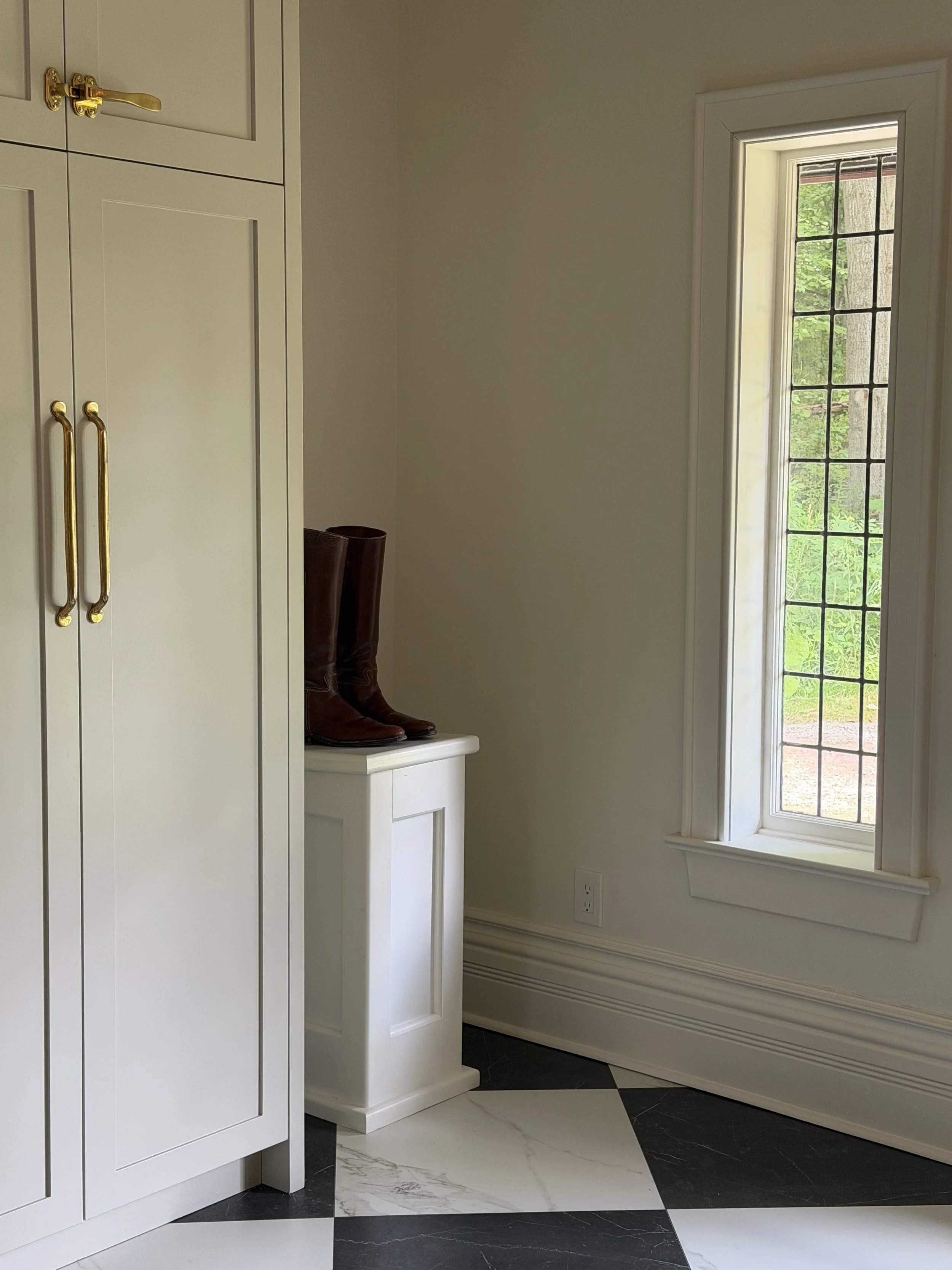 Corner of a room with a white cabinet, a small white stand with brown boots on top, and a large window with divided panes showing greenery outside.
