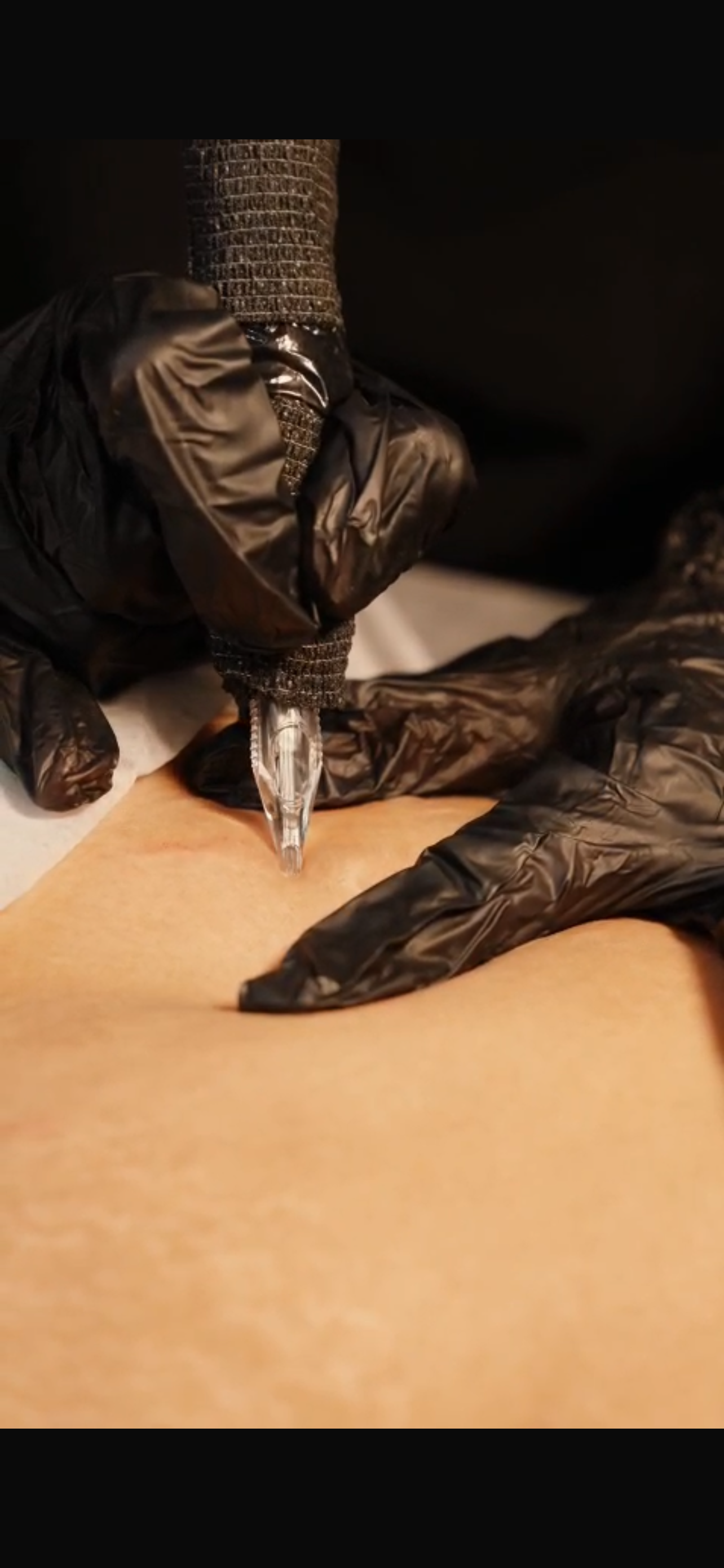 A tattoo artist is tattooing a person’s skin with a tattoo machine. The artist is wearing black gloves and a black protective sleeve.