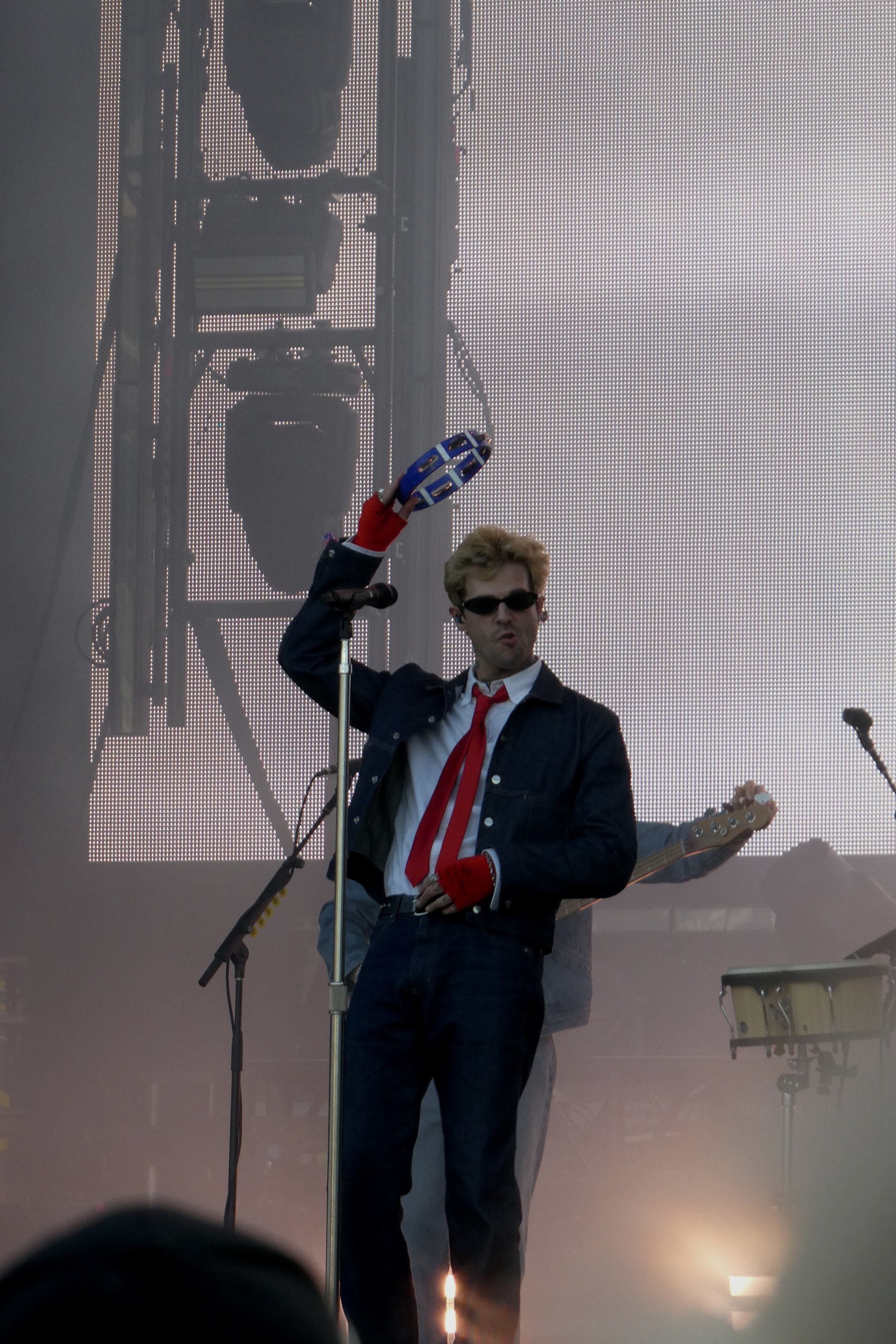 A man in sunglasses, a black jacket, and a white shirt with a red tie is holding a blue tambourine on stage during a concert with a large LED screen and musical instruments in the background.