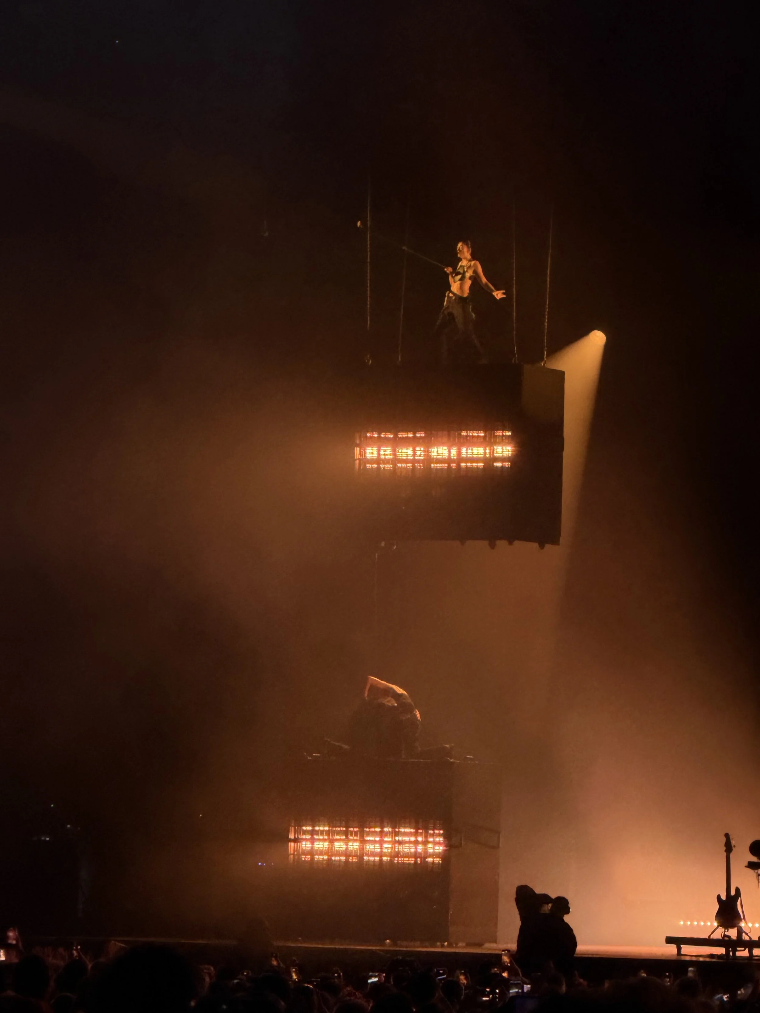 A person standing on stage during a performance, illuminated by orange and yellow lights, with an audience watching and a guitar on a stand to the right.