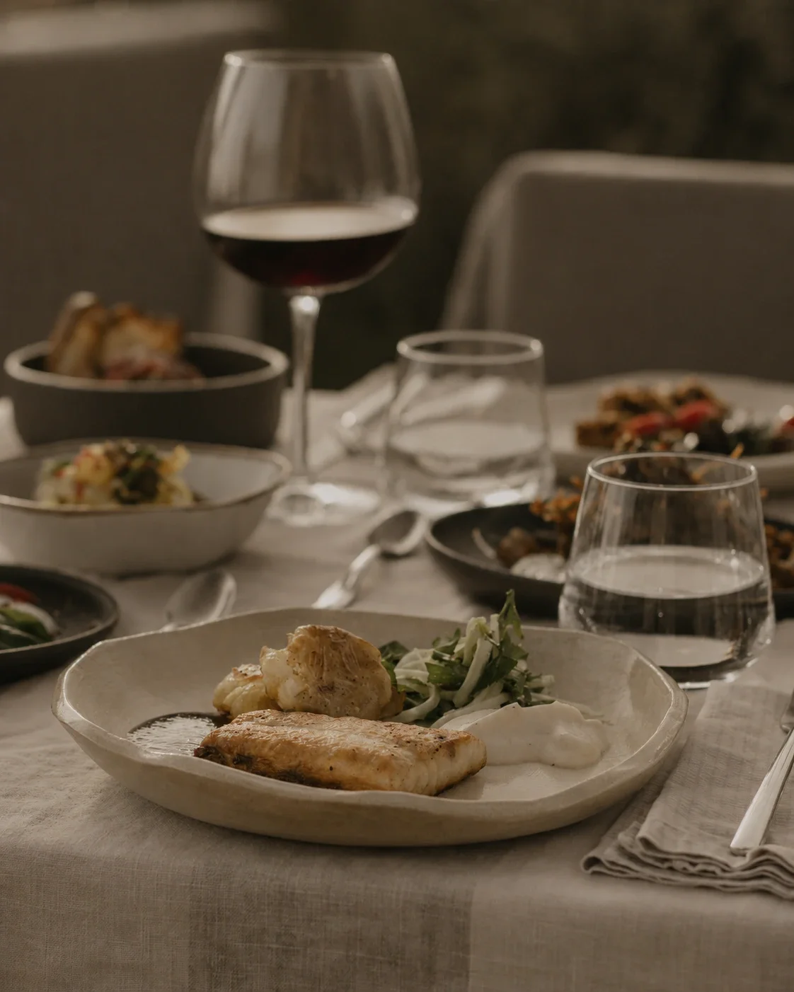 A fine dining table set with a plate featuring fish, cauliflower, and salad, surrounded by glasses of water, a glass of red wine, and various dishes in the background.