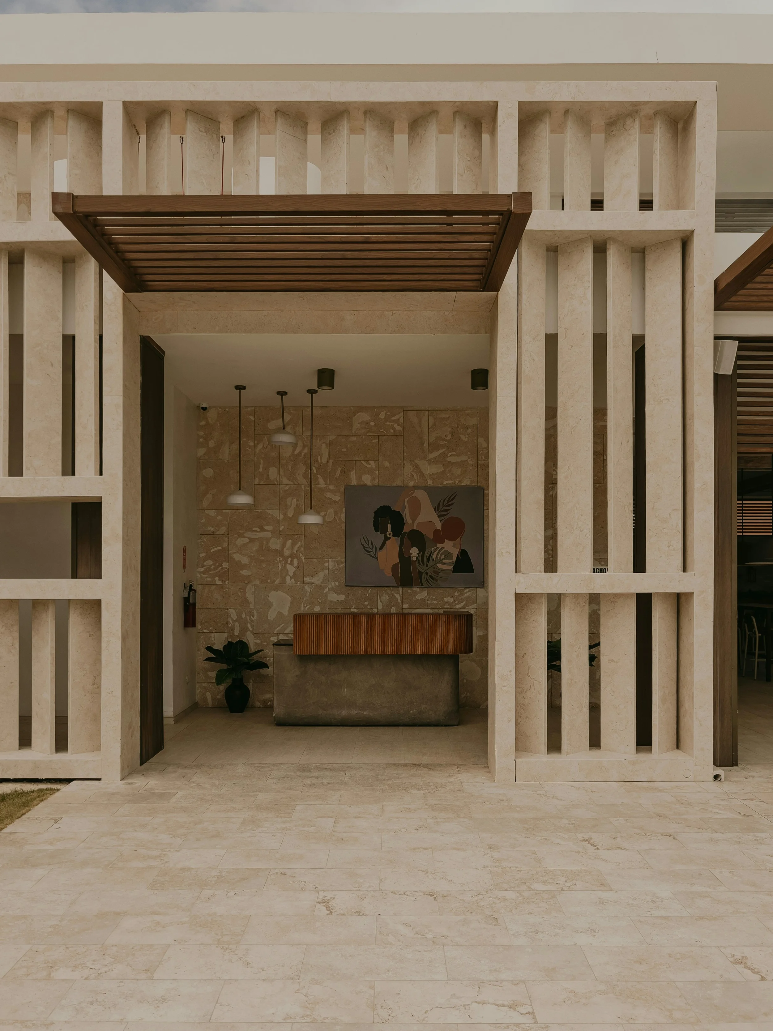 Modern building facade with beige stone walls, vertical stone slats, a covered entrance area with hanging light fixtures, and a piece of abstract artwork on the back wall featuring a woman with natural hair, surrounded by plants and neutral tones.