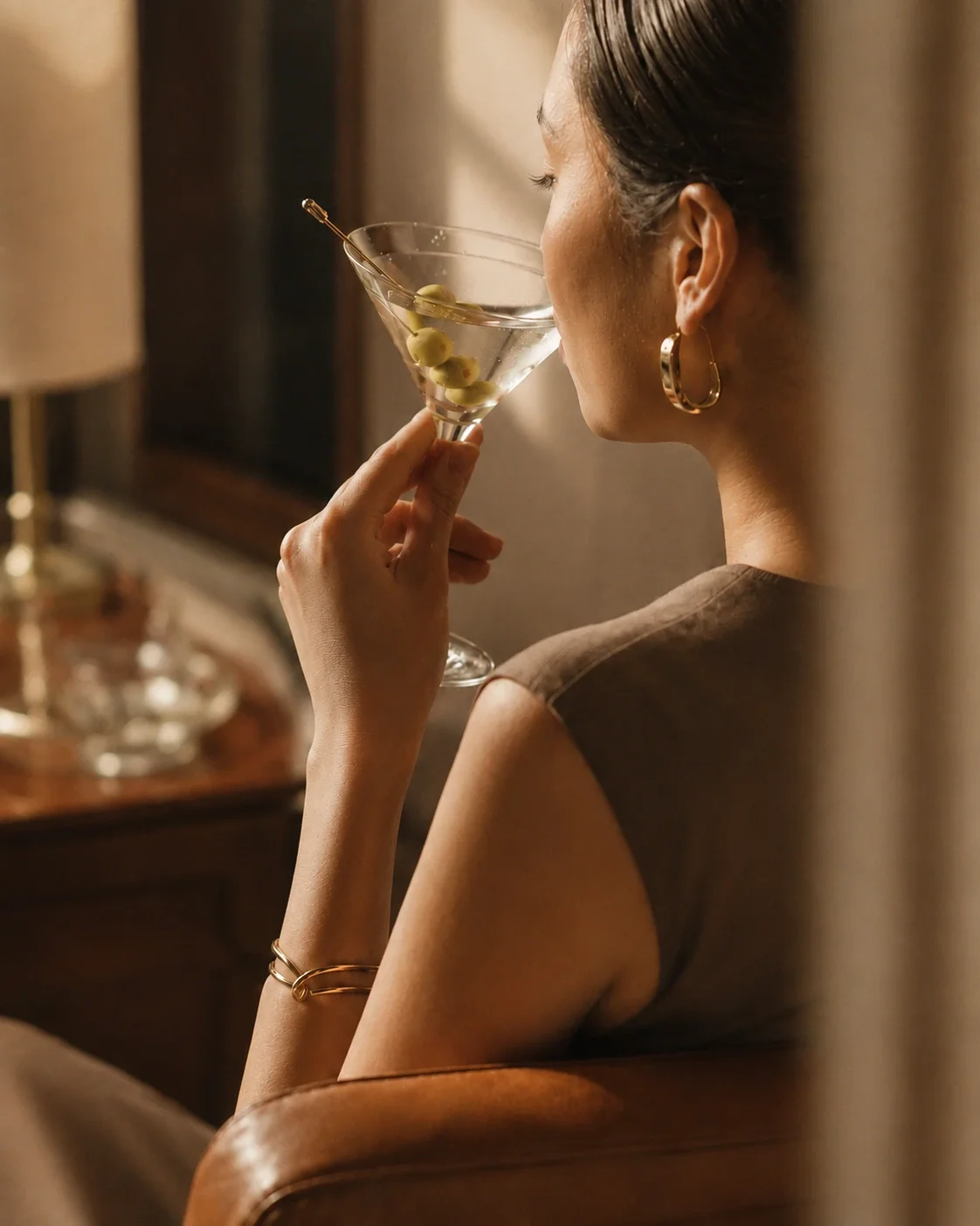 A woman with dark hair and gold hoop earrings sipping a clear cocktail with green olives in a martini glass in a warmly lit room.