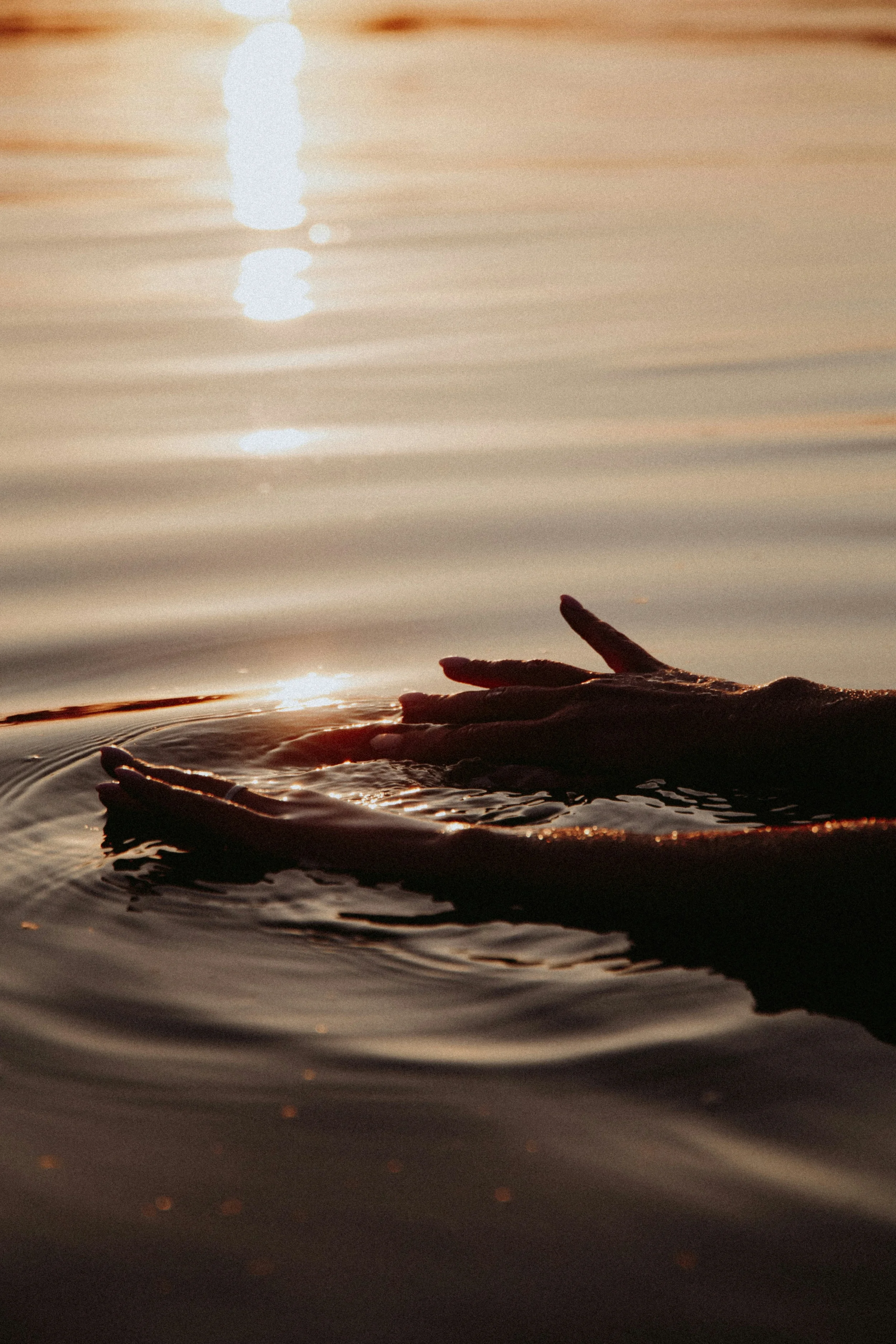 Hands reaching out of calm water during sunset, reflecting warm light on the water's surface.