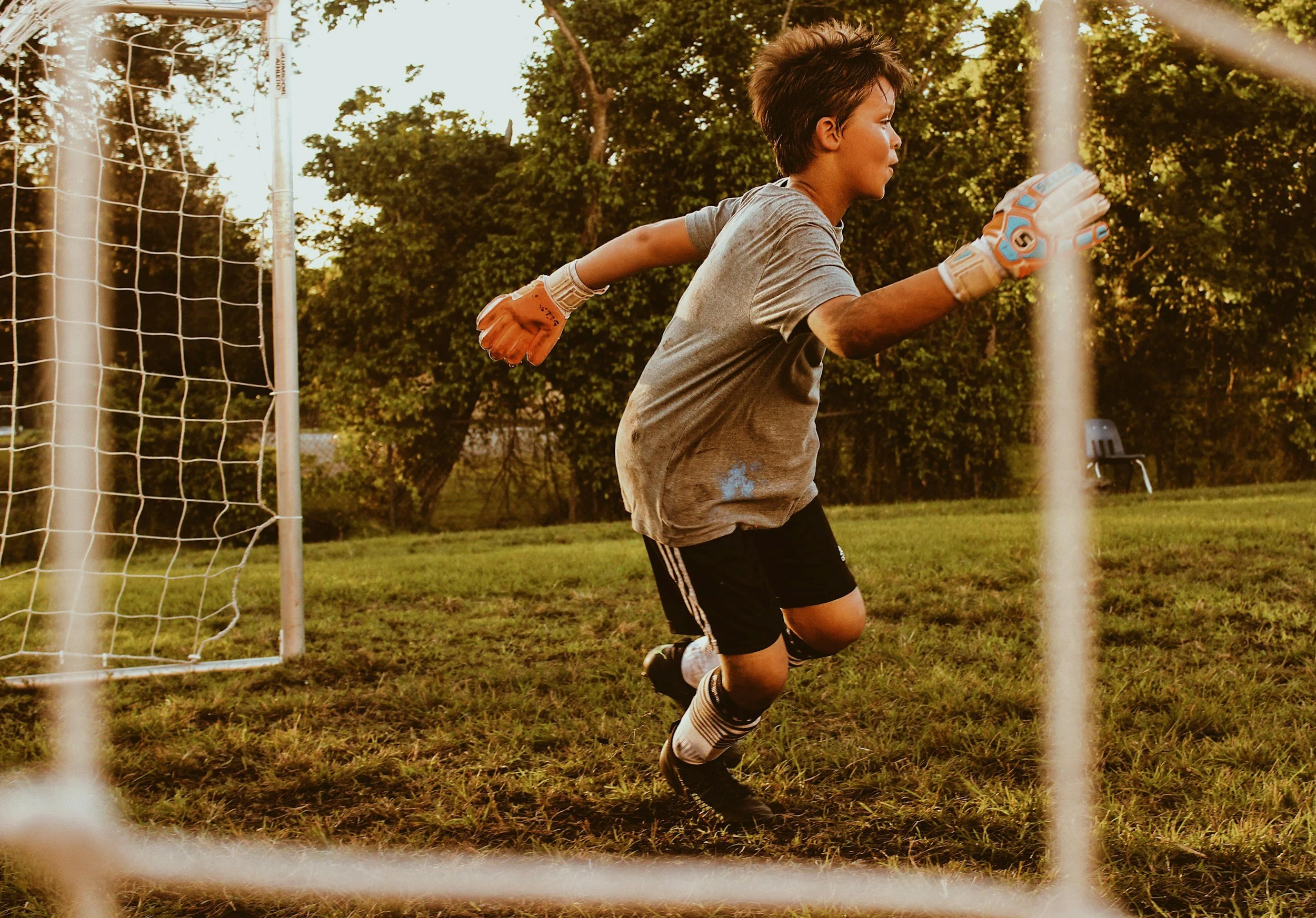 Young boy playing soccer as goalkeeper, wearing gloves, running near the goalpost on a grassy field during sunset, with trees in the background.