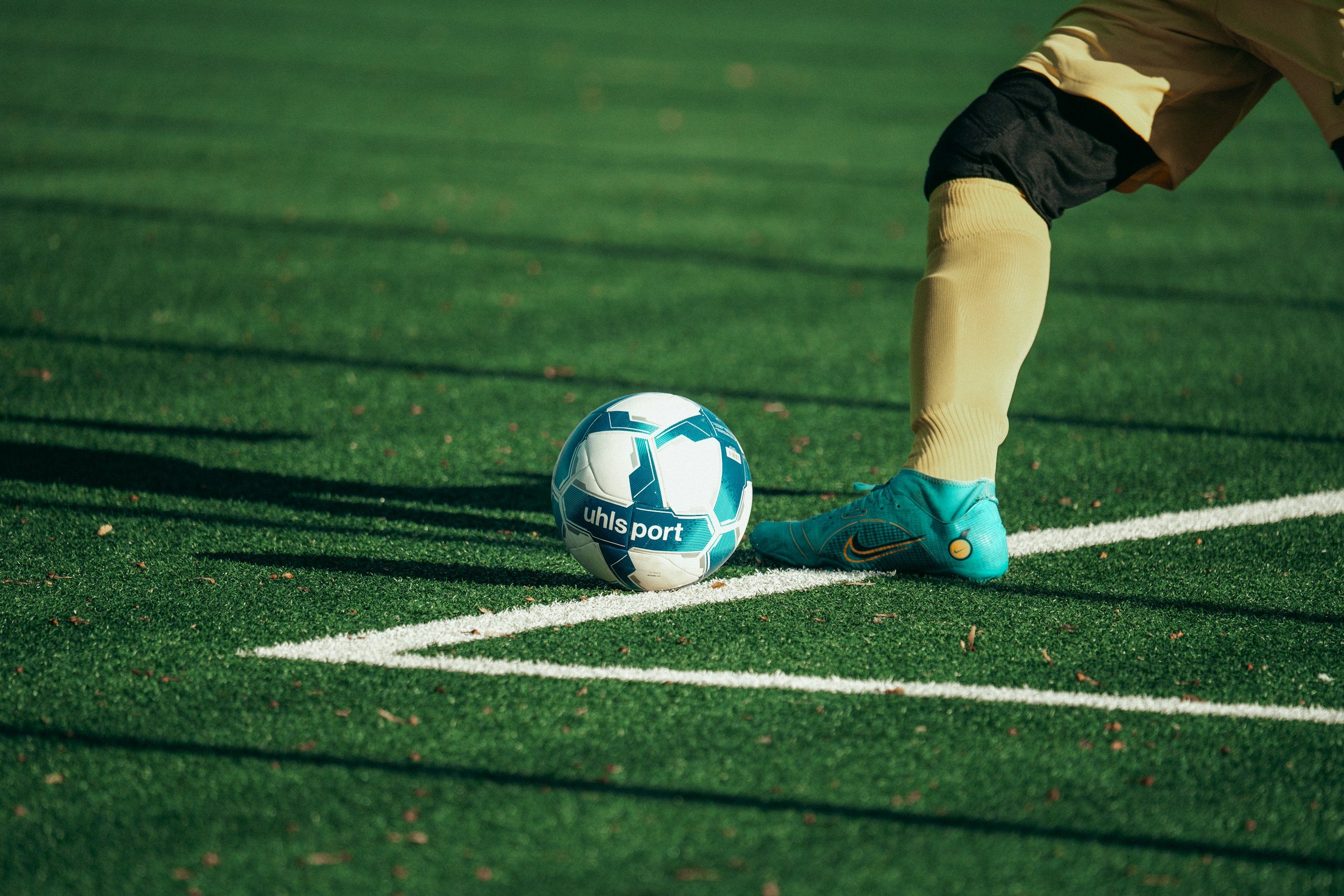 A child in yellow shorts, teal shoes, and navy socks is preparing to kick a white and blue soccer ball on a green field.