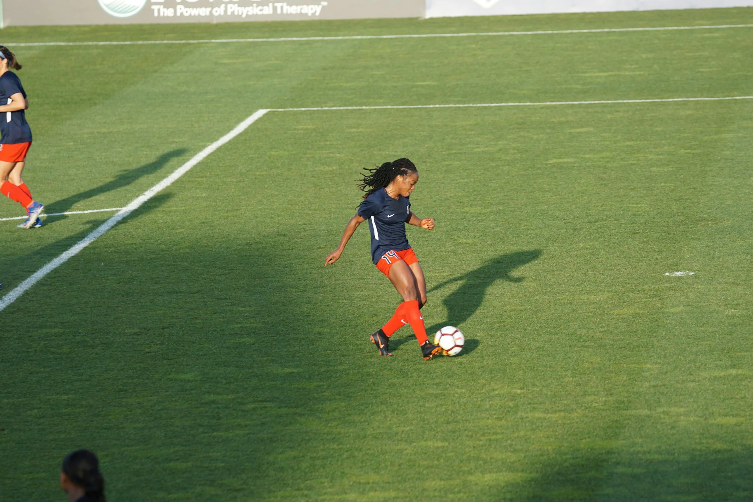 A female soccer player with long braided hair wearing a navy blue jersey and orange shorts dribbling a soccer ball on a green field.