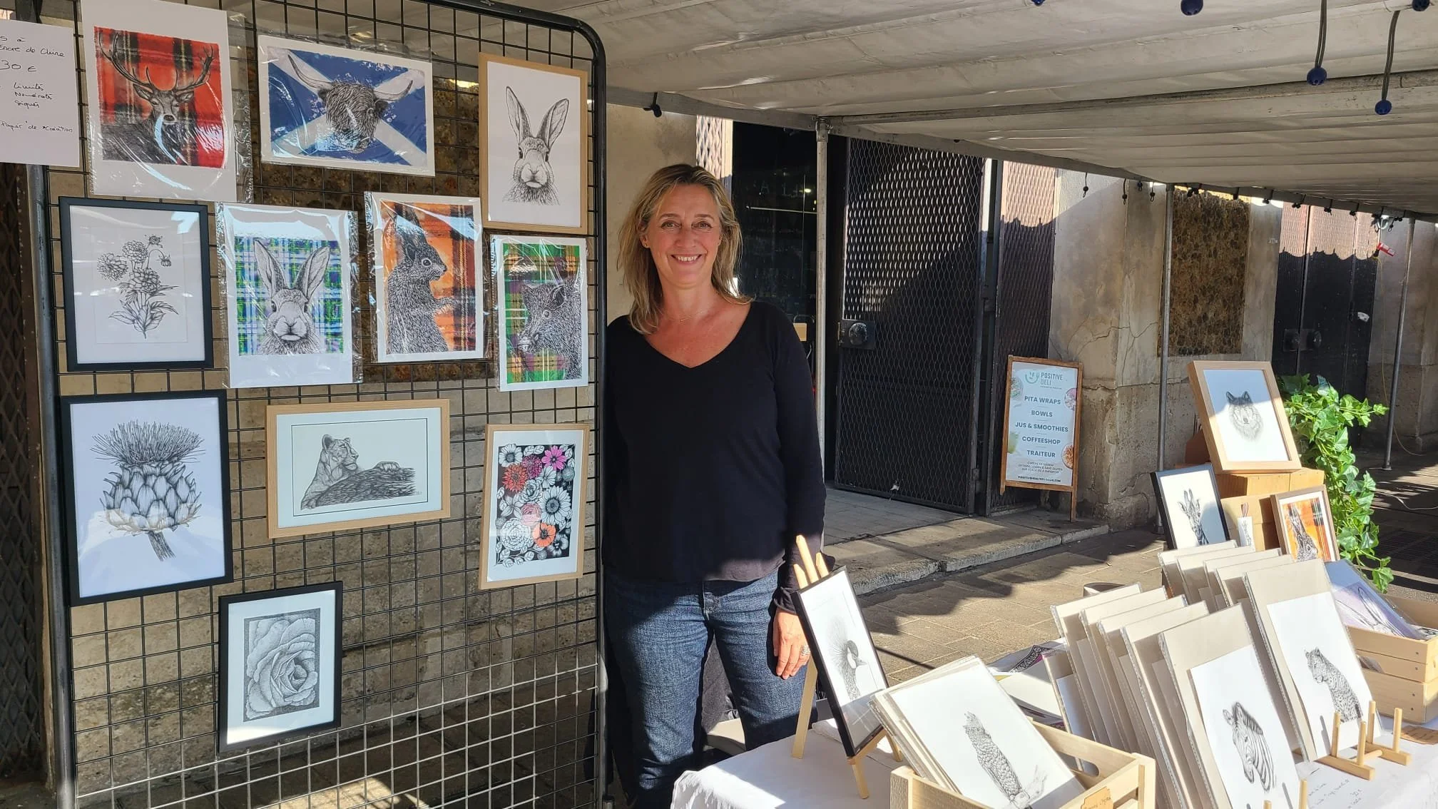 Une femme souriante posant devant un stand d'arts avec des dessins de lapins, de cerfs et d'autres animaux exposés, en plein air.