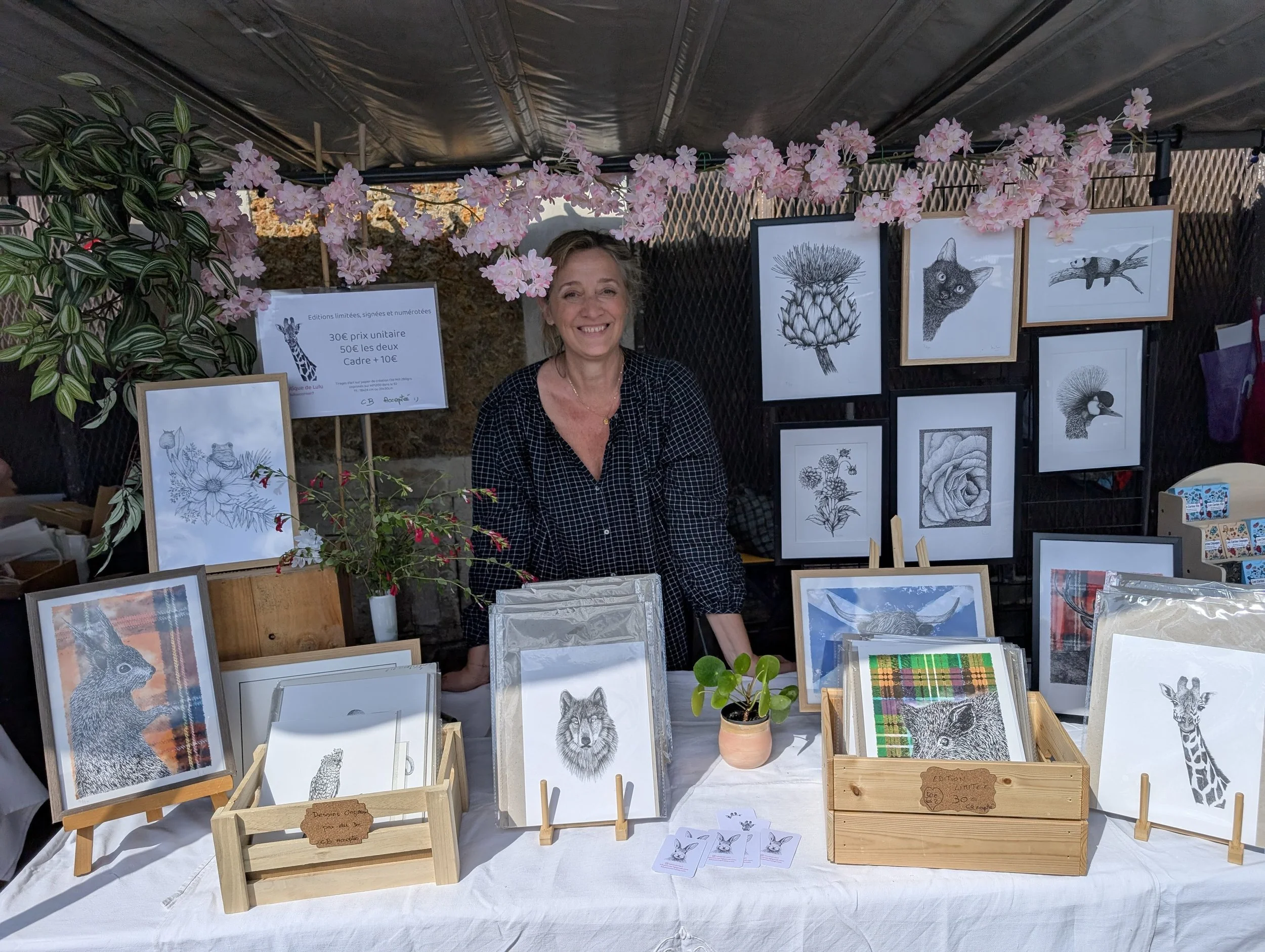 Une femme souriante vendant des œuvres d'art sur un stand lors d'un marché, avec des illustrations de animaux et de plantes encadrées affichées derrière elle, décoré avec des fleurs roses suspendues au plafond.