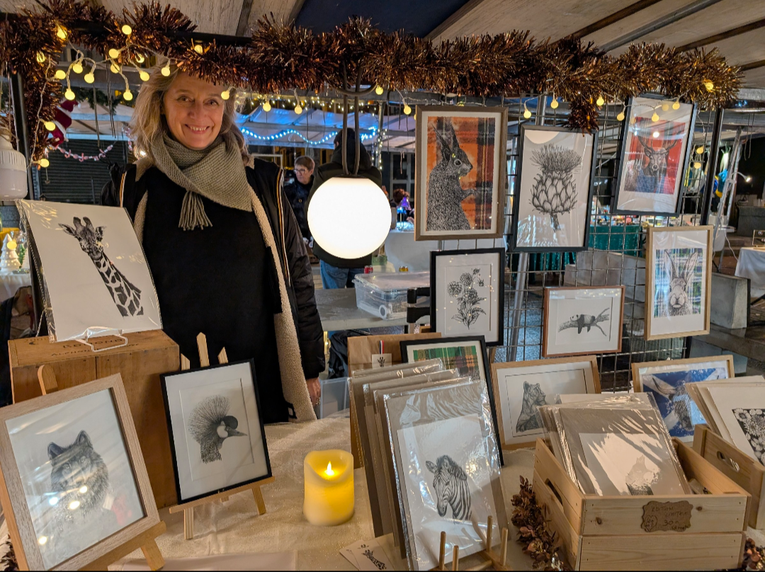 Une femme souriante vend des illustrations d'animaux dans un marché de Noël, avec des portraits d'animaux comme girafe, renard, cerf, et zèbre, décoré de guirlandes lumineuses.