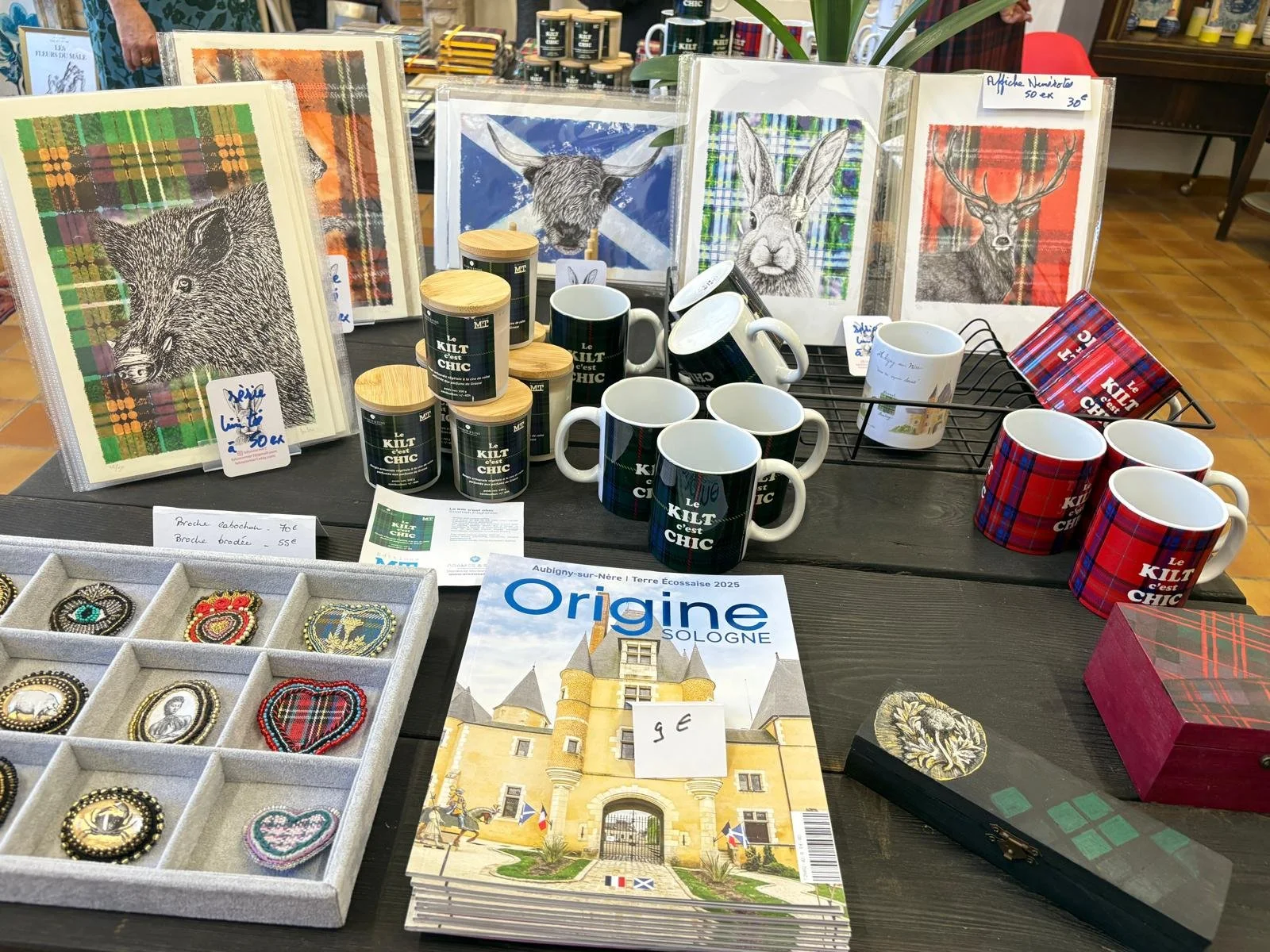 Vente d'articles avec des mugs, des cadres avec des dessins d'un sanglier d'un écureuil, d'un lapin, d'un cerf et d'une vache écossaise sur fond en tartan. Des broches, et un magazine intitulé 'Origine Sologne' sur un stand dans un magasin.