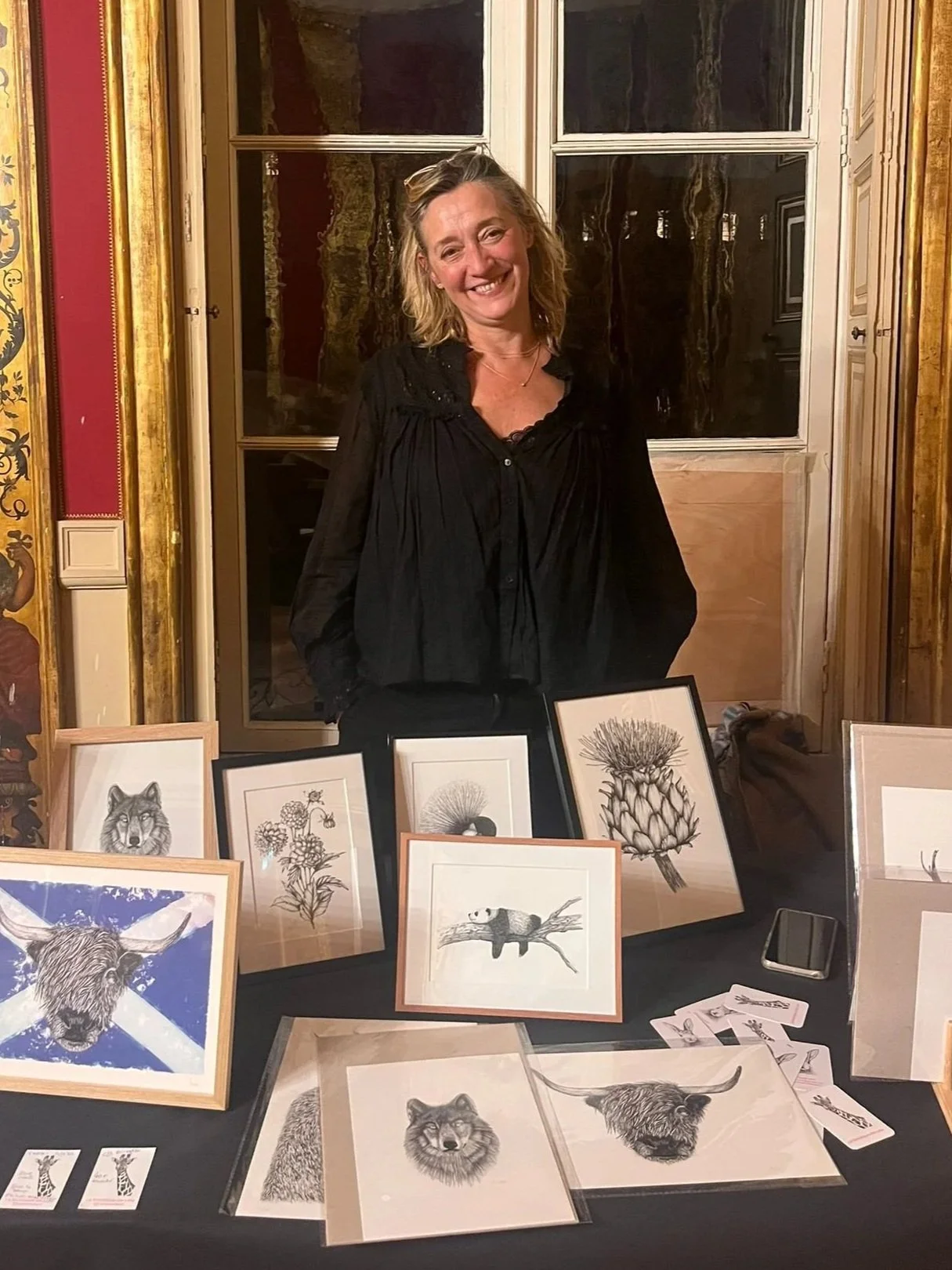 Une femme souriante se tient derrière une table d'exposition avec des dessins d'animaux et de nature, notamment un loup, une bêche, un chardon et un caméléon.
