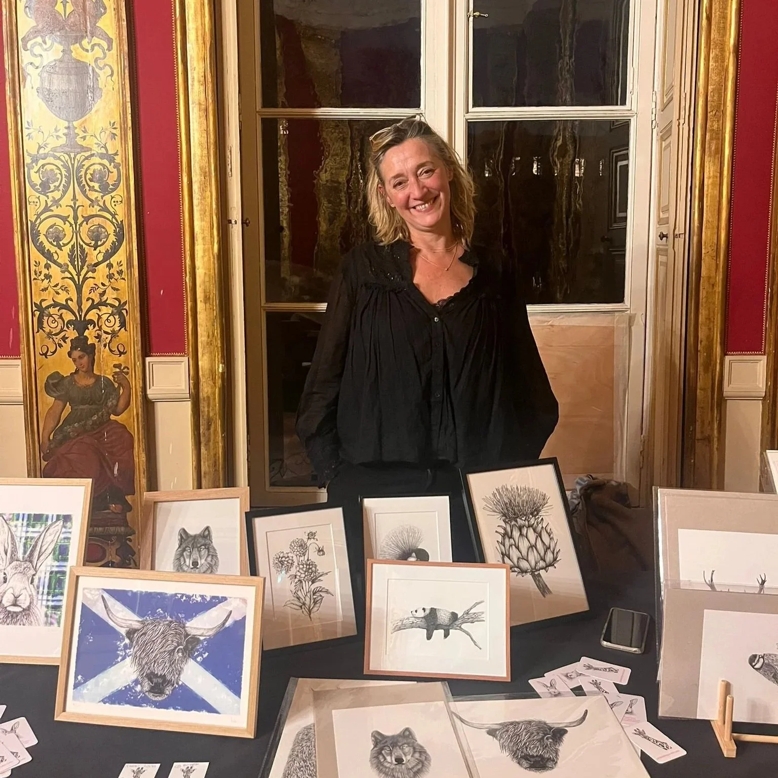Une femme souriante se tient derrière une table d'exposition avec plusieurs dessins d'animaux, notamment un taureau, un chien, un renard et une chouette. La scène semble se dérouler dans un lieu élégant avec des murs ornés de dorures et des tableaux ou décorations anciennes en arrière-plan.