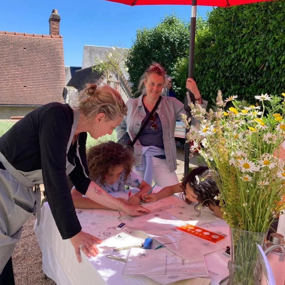 Deux femmes aidant des jeunes à dessiner ou colorier sur une table lors d'un atelier en plein air, avec un bouquet de fleurs sauvages en premier plan et un soleil éclatant.