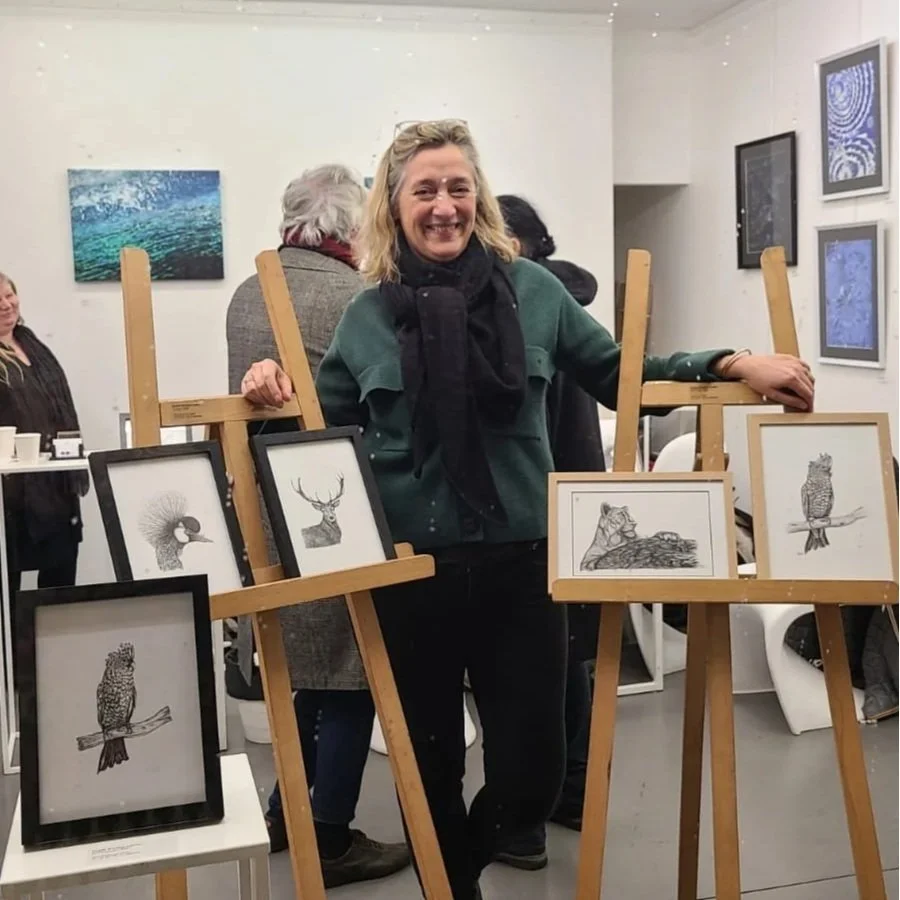 Une femme souriante posant dans une galerie d'art, entourée de dessins encadrés représentant des animaux, notamment un paon, un cerf, un lion, un hibou, et un hibou perchoir.