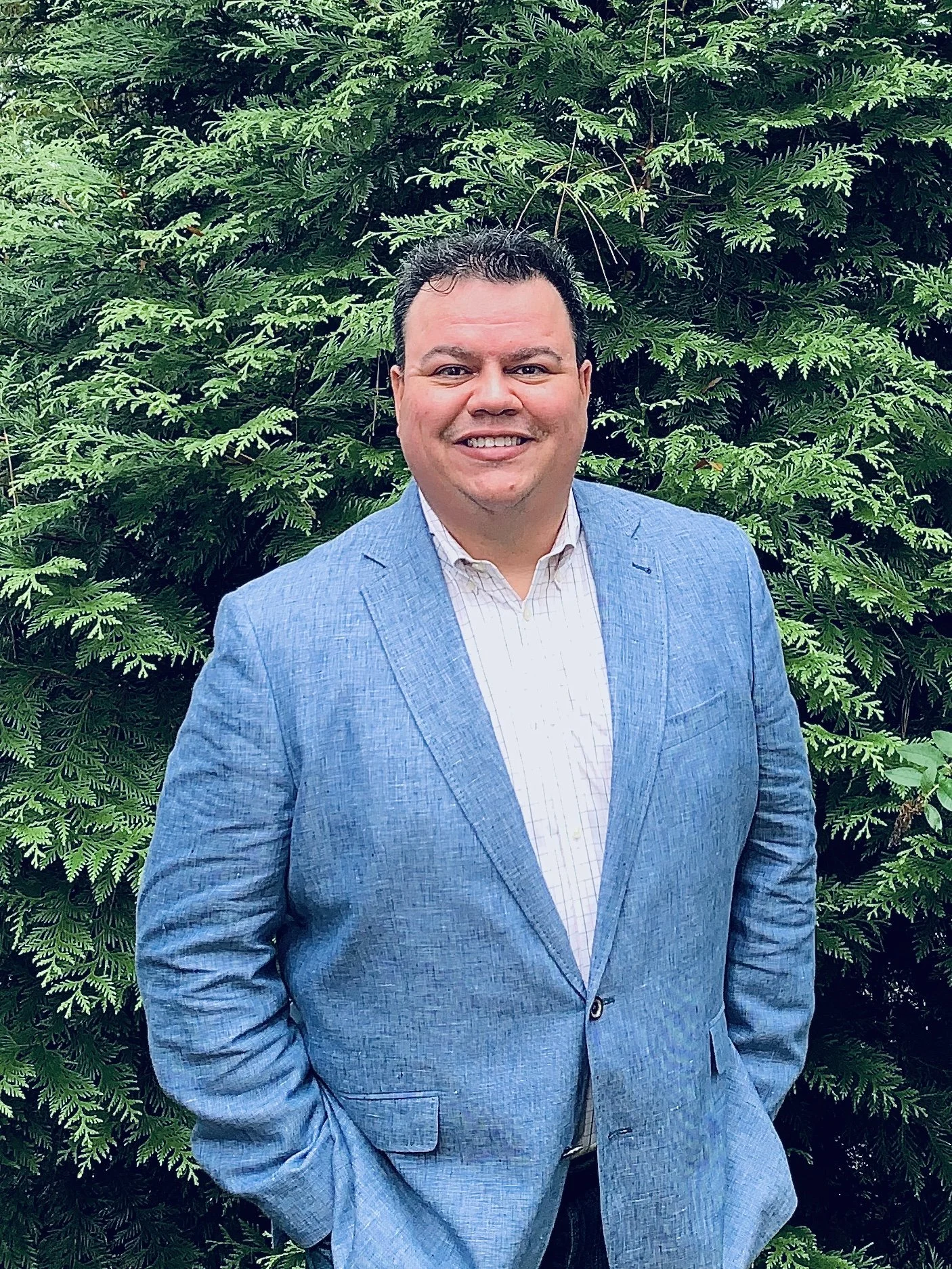 A smiling man in a blue suit jacket and white dress shirt standing outdoors in front of green ferns and foliage.