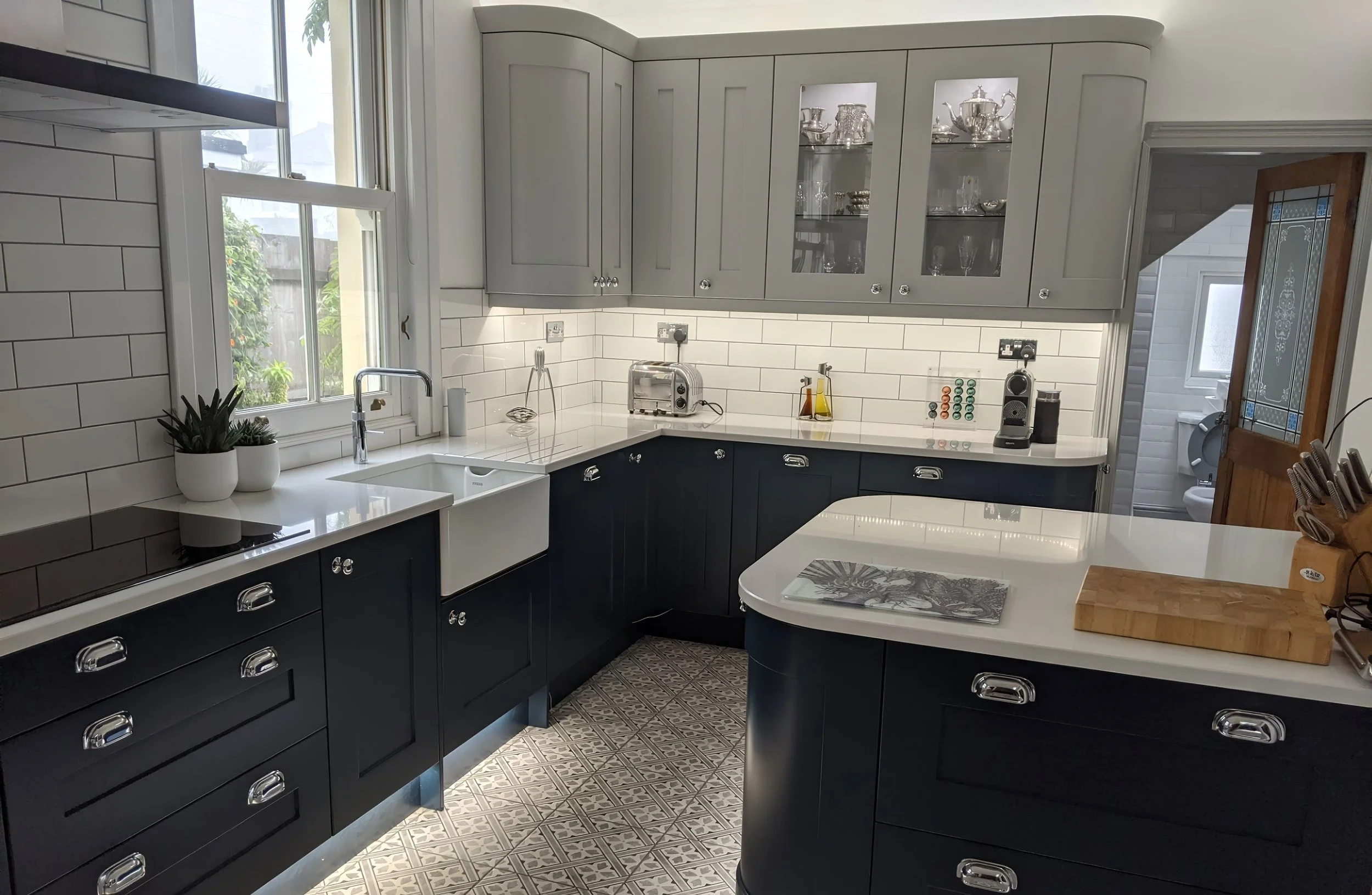 Modern kitchen with white cabinets, black lower cabinets, white subway tile backsplash, large window, and patterned tile floor. Countertop items include a plant, toaster, oil bottles, coffee machine, and paper towel roll.