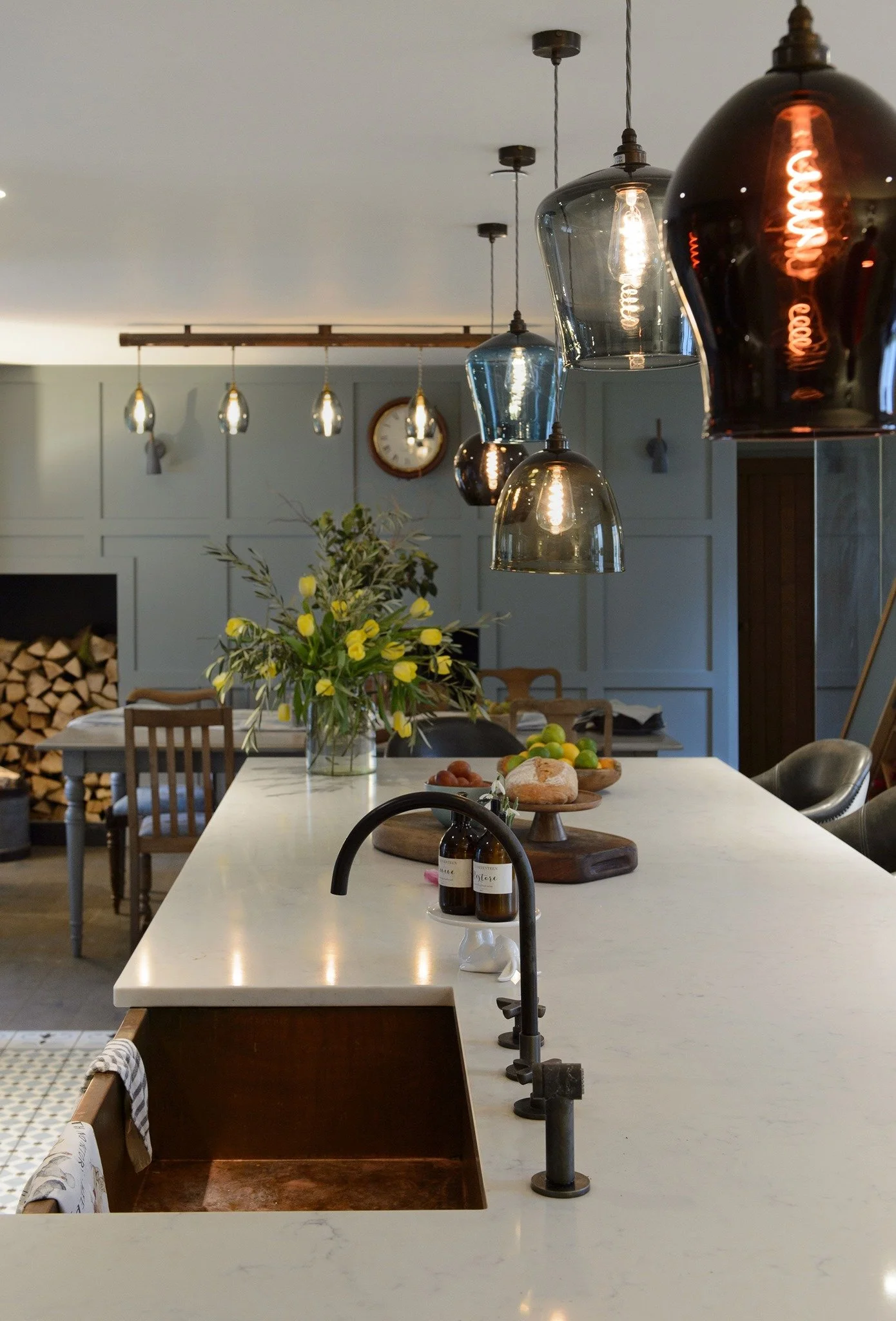 Modern kitchen or dining area with a white marble island countertop, black and gold pendant lights hanging from the ceiling, a bouquet of yellow flowers and fruit on the counter, and a wall clock in the background. The room has a cozy, stylish atmosphere.
