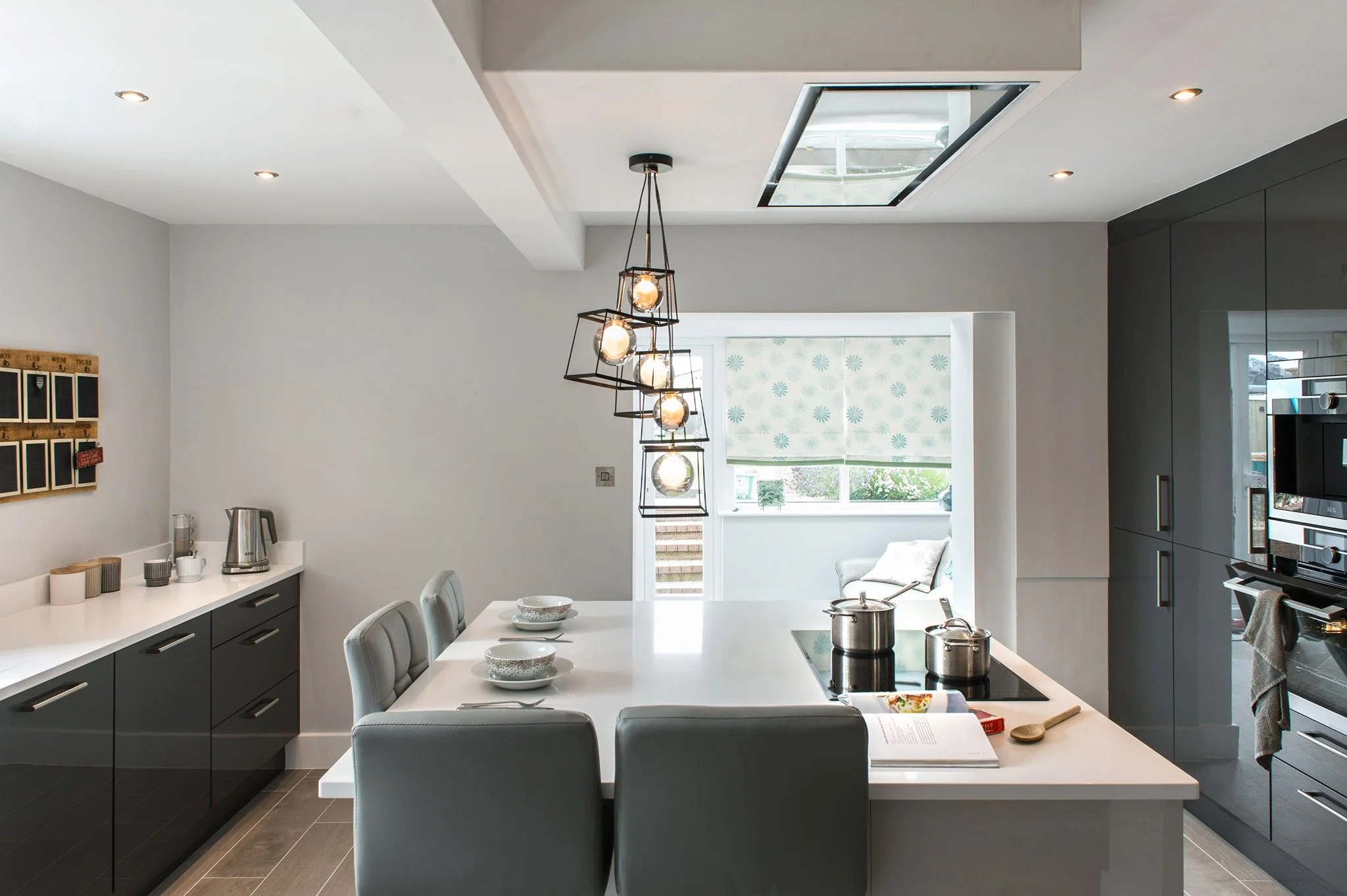 Modern kitchen with white island counter, gray cabinets, and dining table with four chairs. Hanging pendant lights above the table, large window, and a skylight in the ceiling.