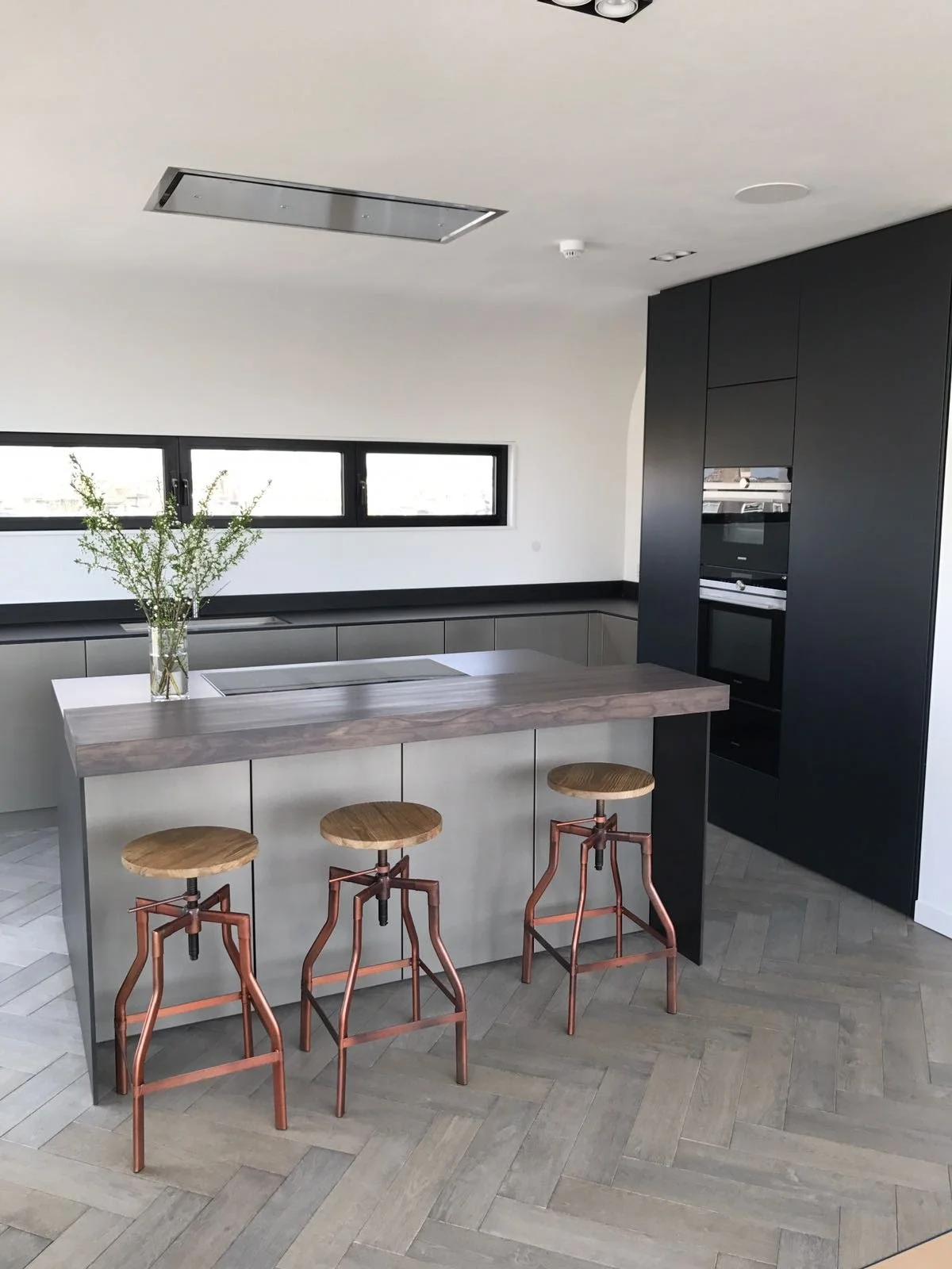 Modern kitchen with black cabinets, a wooden kitchen island with three copper and wooden stools, a black window, and minimal decor with a vase of green branches.