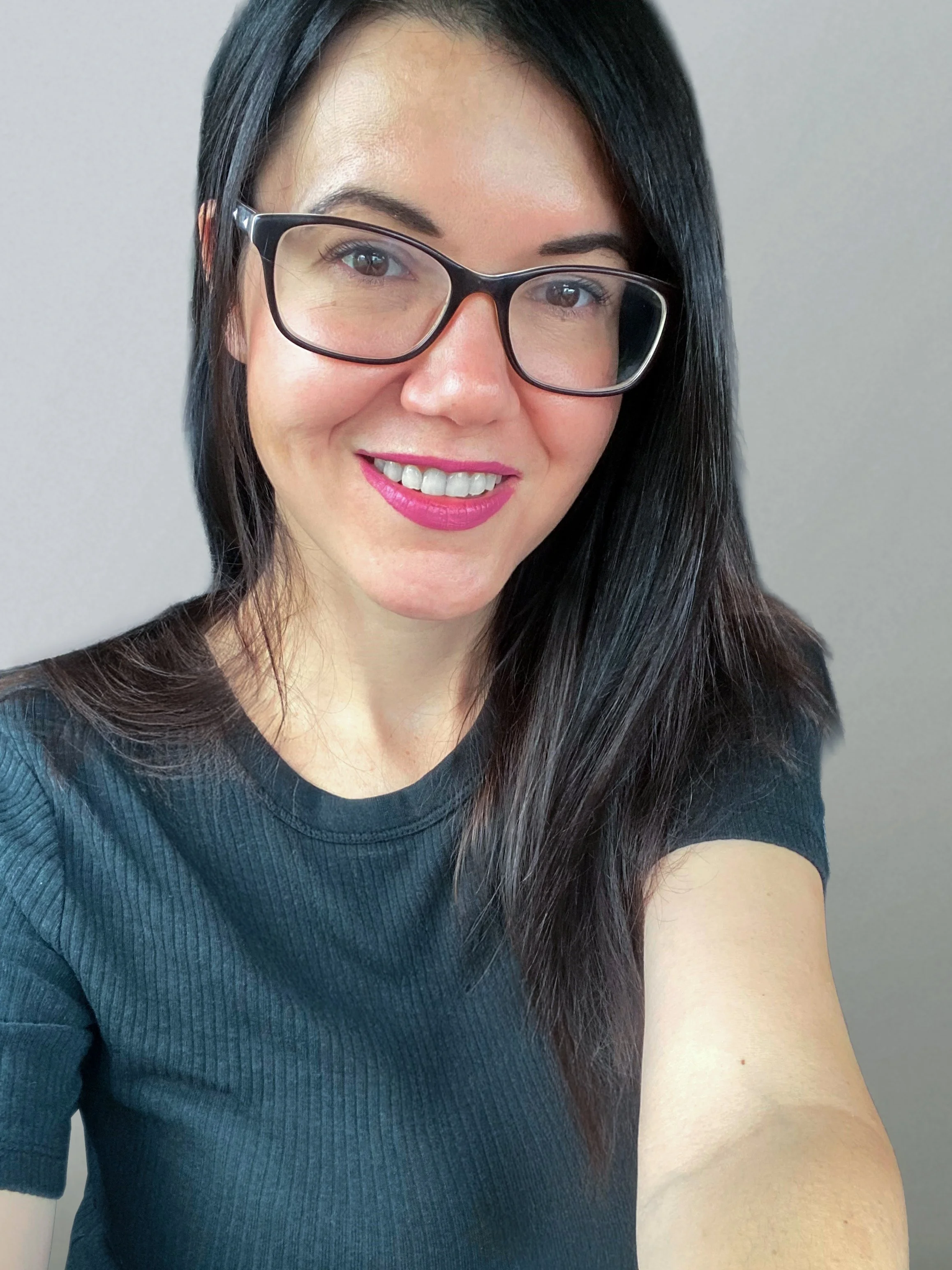 A woman with long black hair, glasses, and pink lipstick, smiling in a close-up selfie against a gray background.