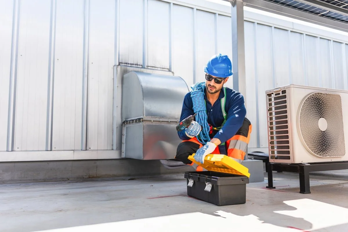 A worker wearing a blue hard hat, sunglasses, and safety gear is kneeling on a rooftop, using a tool from a yellow toolbox.