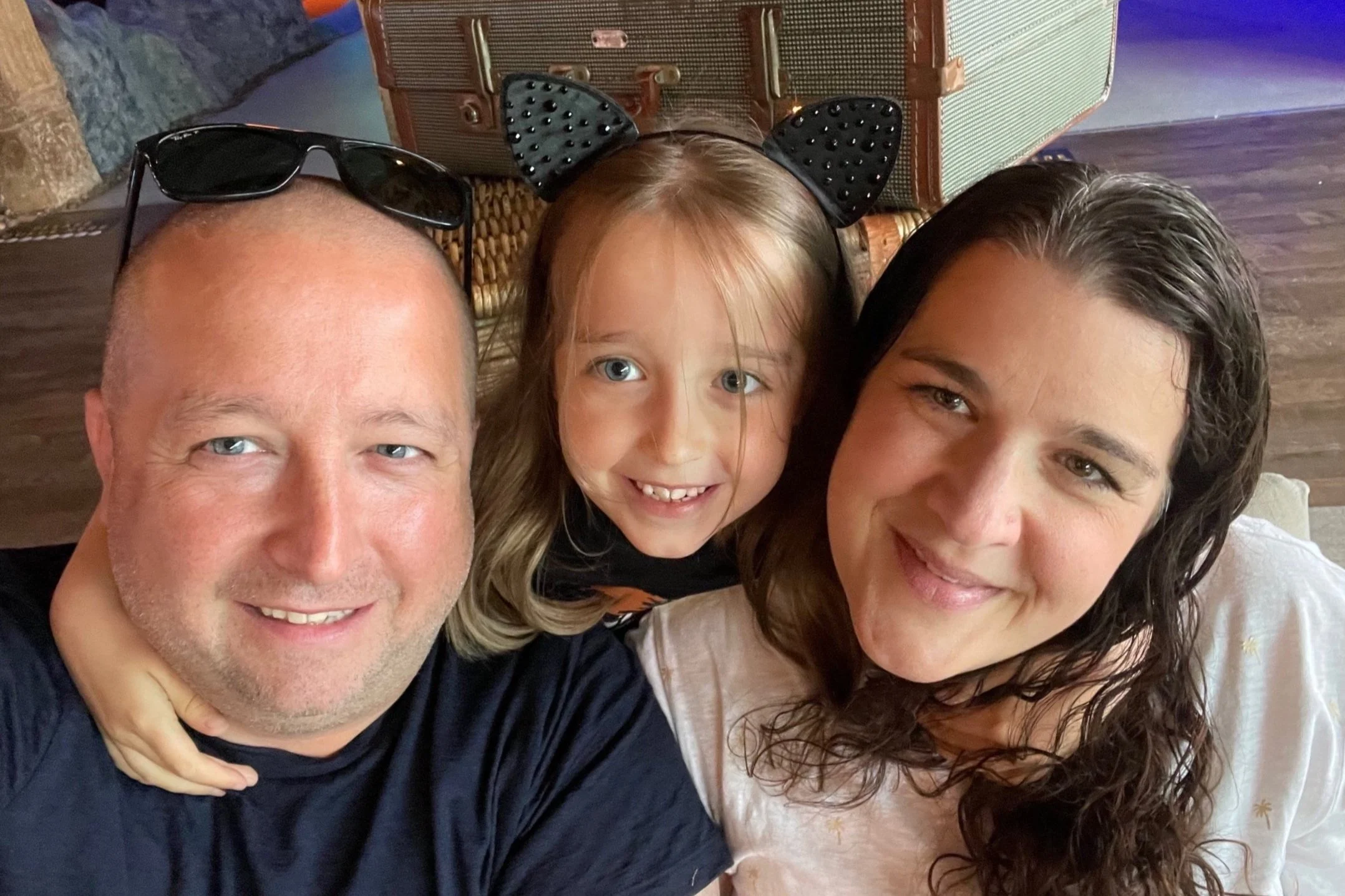 Close-up photo of three smiling people, two adults and a young girl, taking a selfie together indoors. The man on the left has short hair and sunglasses on his head, the young girl in the middle has long hair and is wearing cat ear headbands, and the woman on the right has curly dark hair.