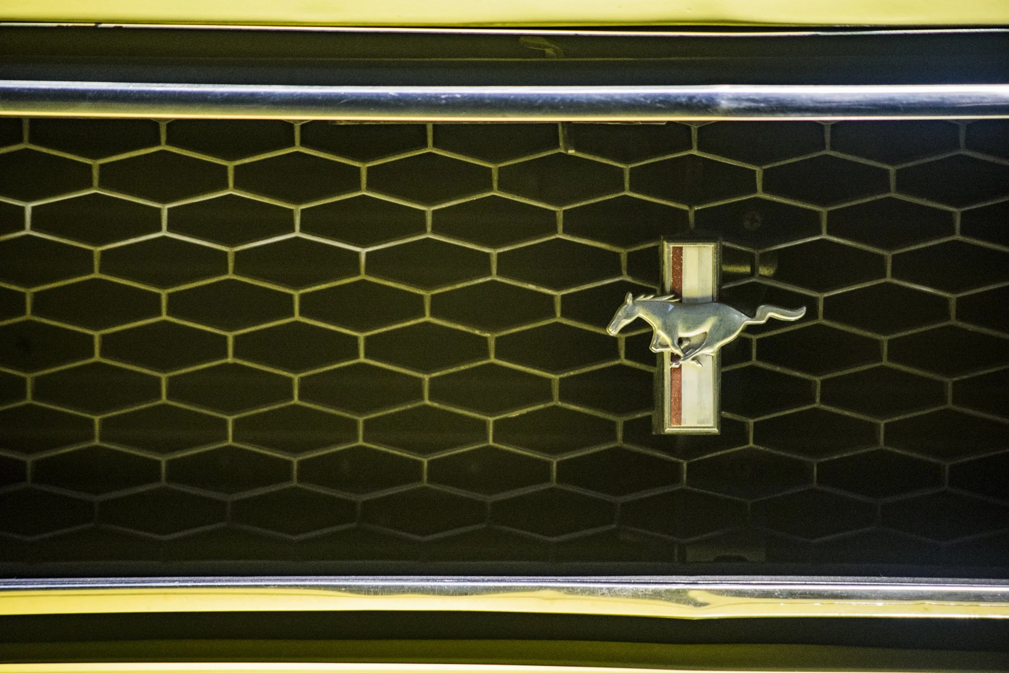 Close-up of a black honeycomb grille with a silver and black Mustang emblem in the center, part of a yellow car.