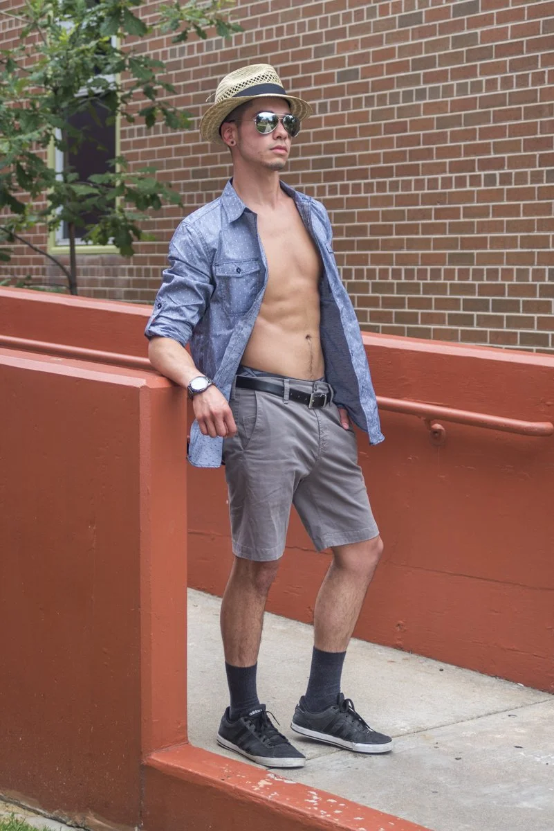 A young man with sunglasses and a straw hat, shirt open showing his torso, standing outside against an orange wall with a brick building and some green foliage in the background.