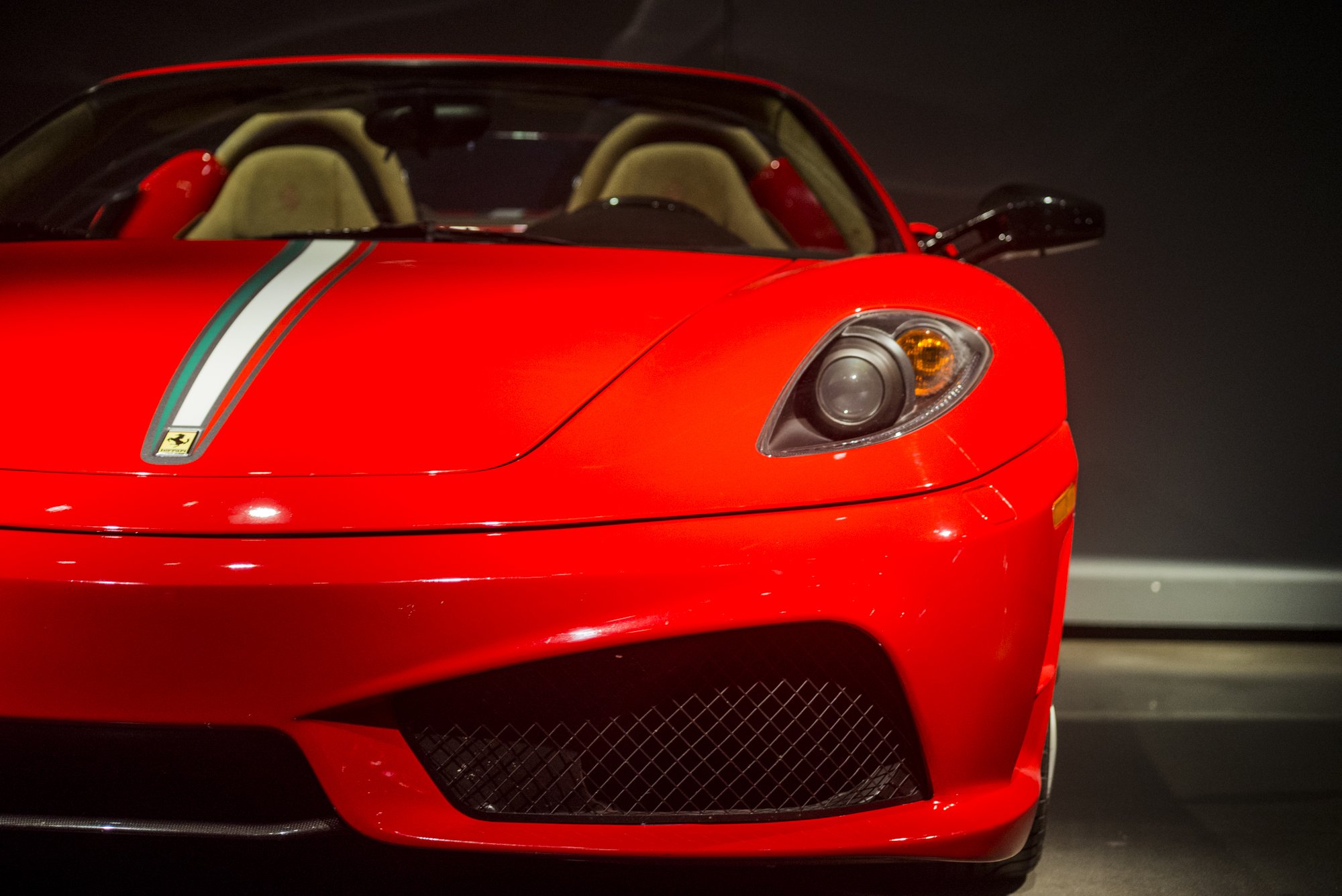 Close-up of the front of a red sports car, likely a Ferrari, showing the headlight, grille, and a racing stripe with Ferrari emblem.