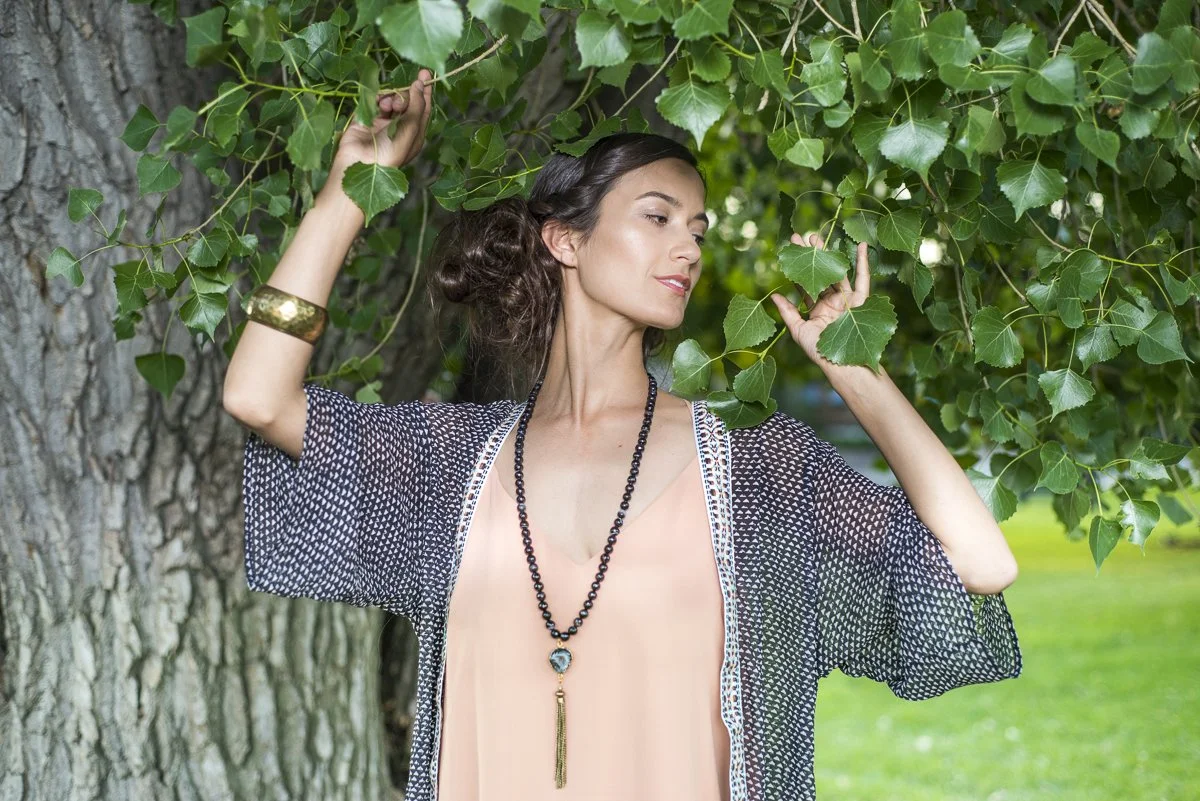 A woman with dark hair and a floral patterned shawl standing outdoors beside a tree, gently touching the leaves with her eyes closed.
