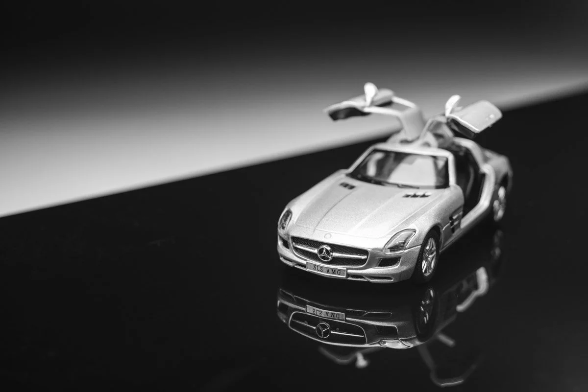 Black and white photo of a miniature Mercedes-Benz SLS AMG toy car with gullwing doors open, placed on a reflective surface.