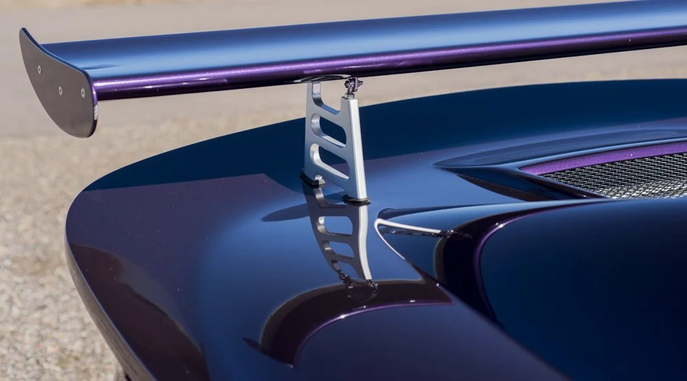 Close-up of a dark blue sports car's front hood with a vertical silver racing wing support and a wing mounted on top.