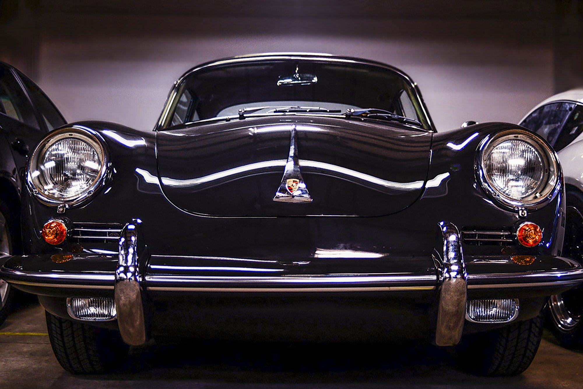 Front view of a black vintage Porsche 356 car displayed indoors, flanked by other cars, with visible headlights, bumper, and Porsche emblem on the hood.