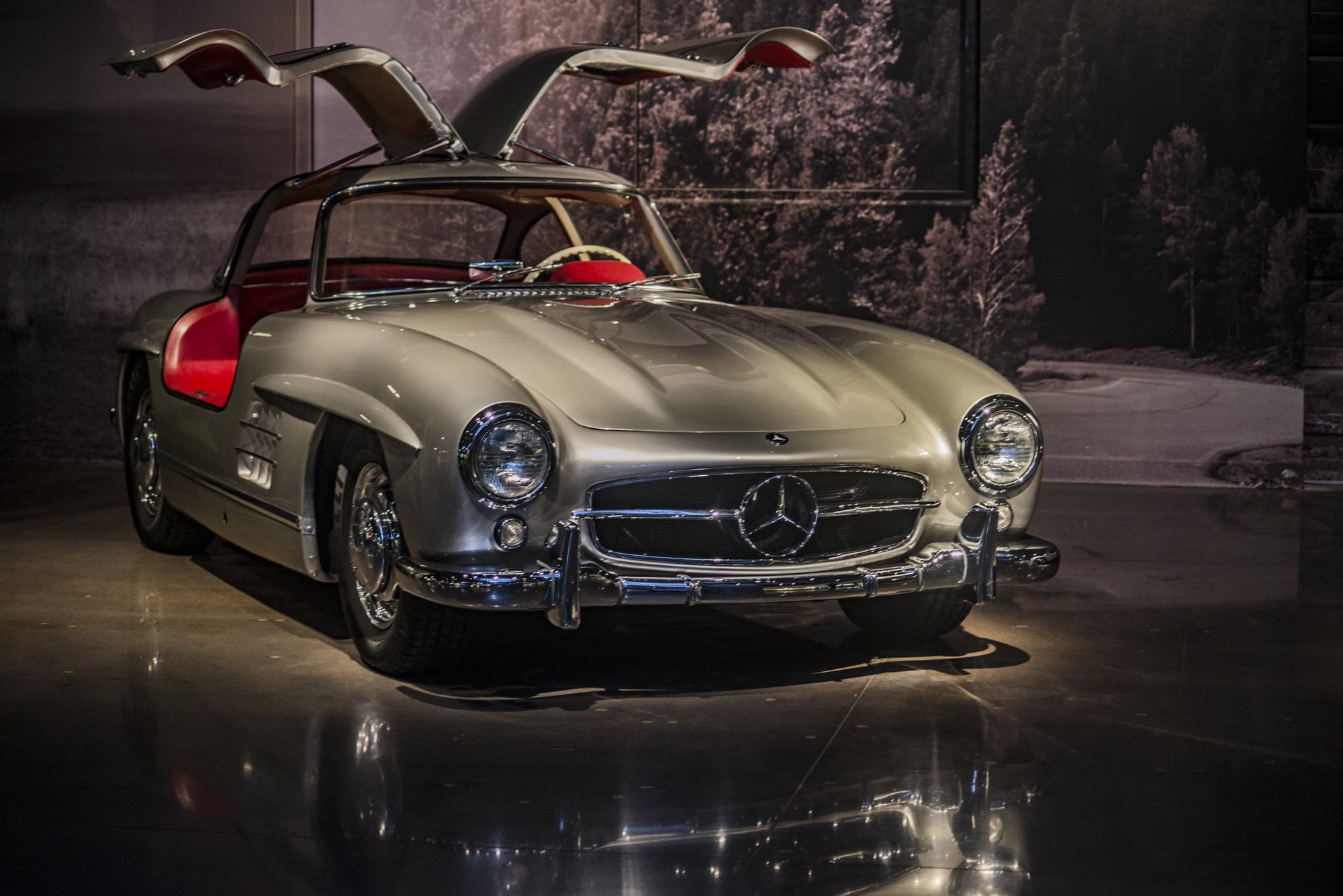 A vintage Mercedes-Benz 300SL gullwing car in a museum setting with a dark background and forest scene backdrop.