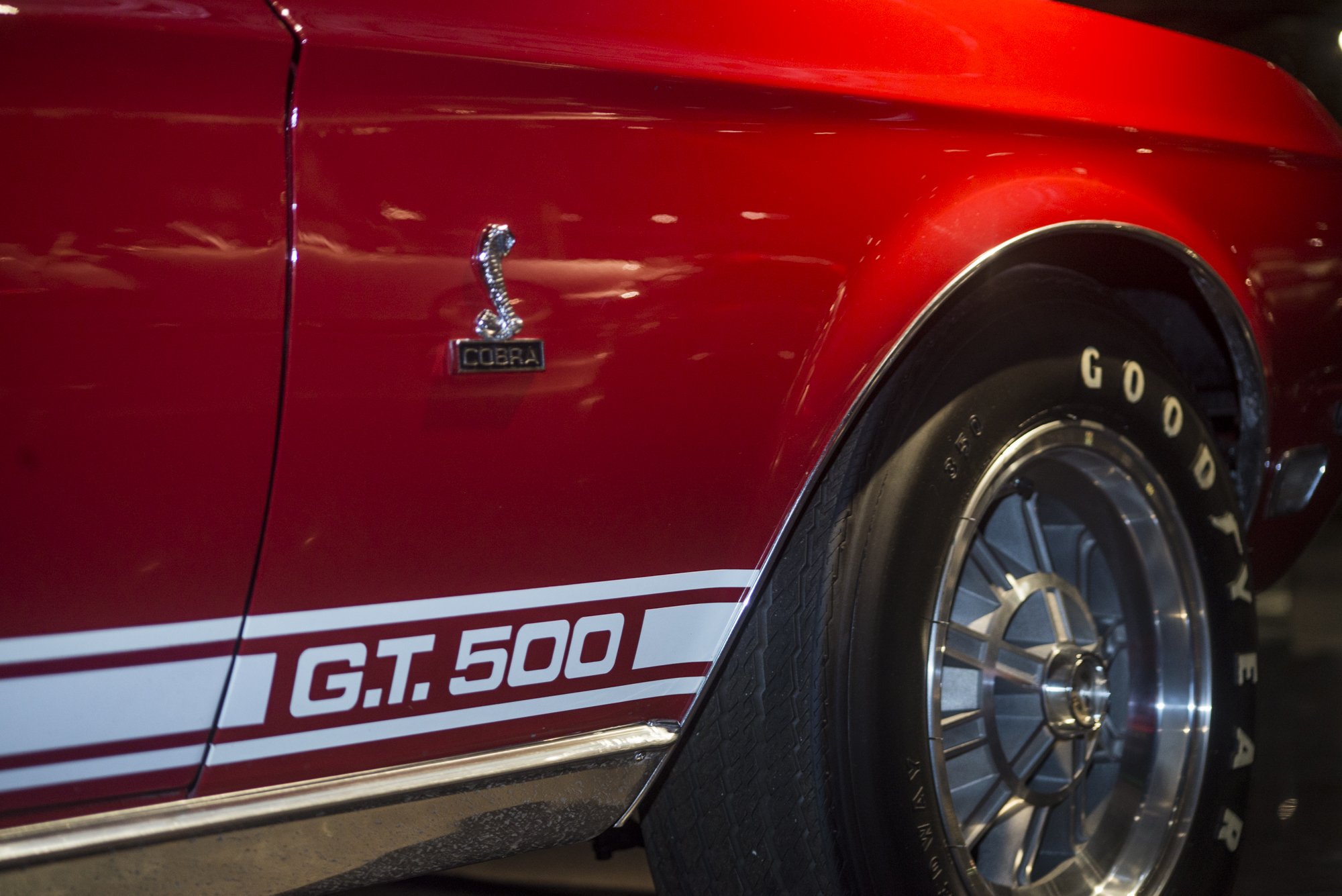 Close-up of a red classic Ford Mustang Cobra driven car, featuring Cobra emblem badge and white racing stripes with 'GT 500' on the side, along with a visible racing tire and wheel.