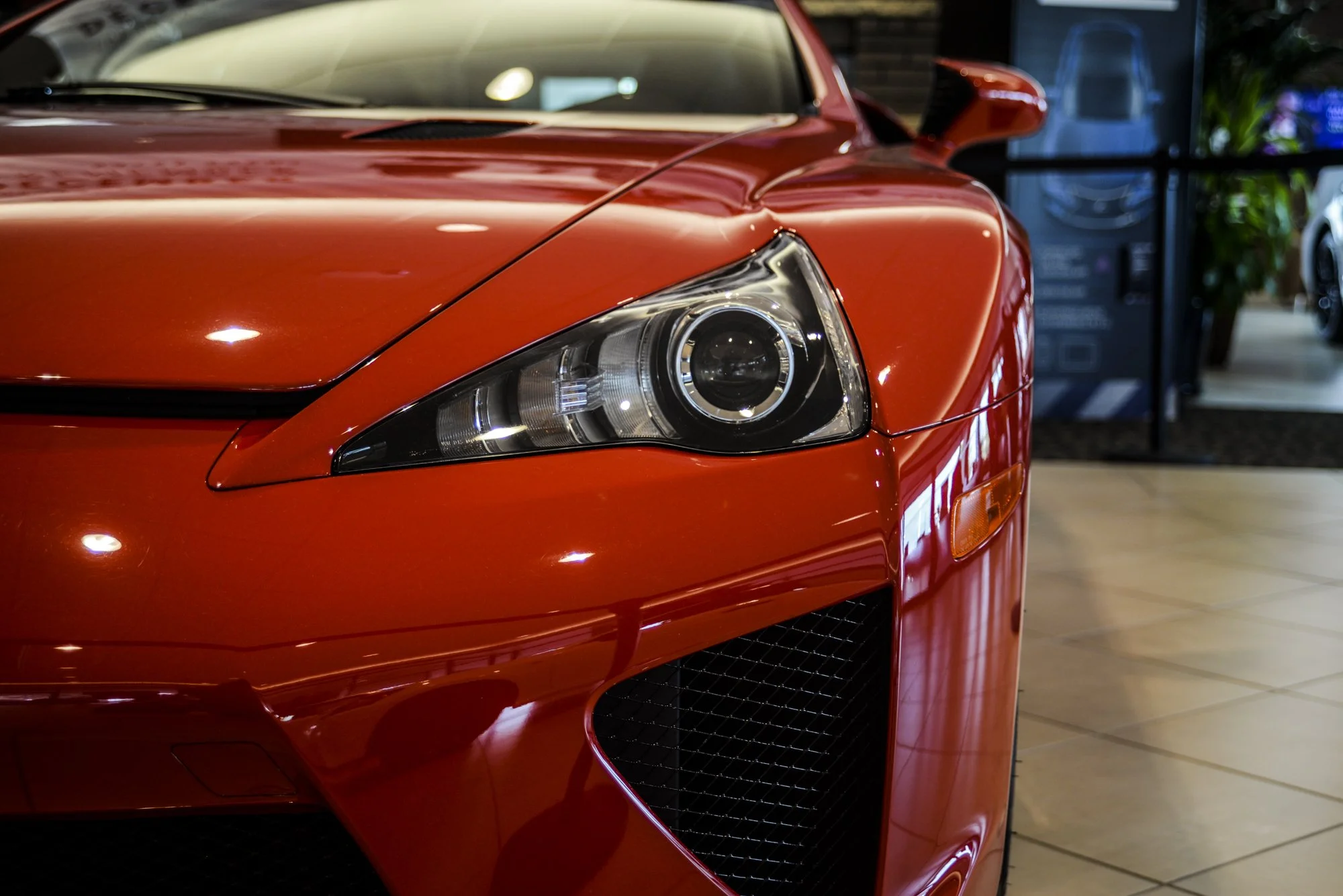 Close-up of the front part of a shiny red sports car, showcasing the headlight, hood, and side mirror, indoors at a dealership.