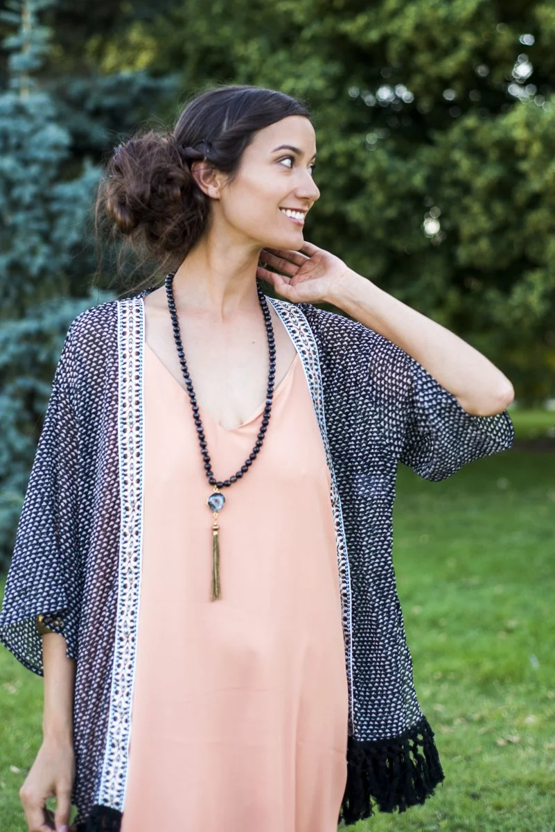 A woman with dark hair styled in a loose bun with a braid, smiling and looking to her right, standing outdoors in a park with green trees and grass in the background. She is wearing a peach dress, a black beaded necklace with a decorative pendant, an
