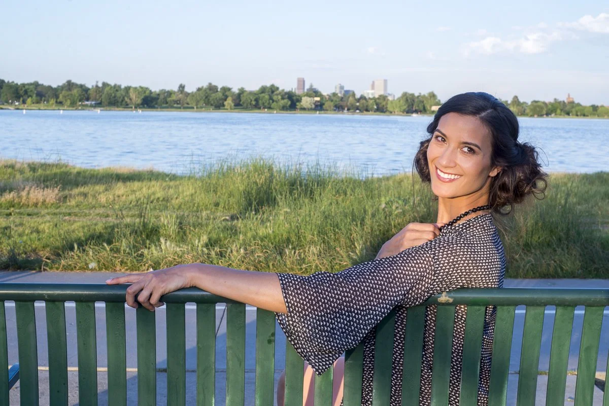 A woman with dark hair and a black and white patterned top smiling and leaning on a green metal railing by a river, with greenery and city buildings in the background.