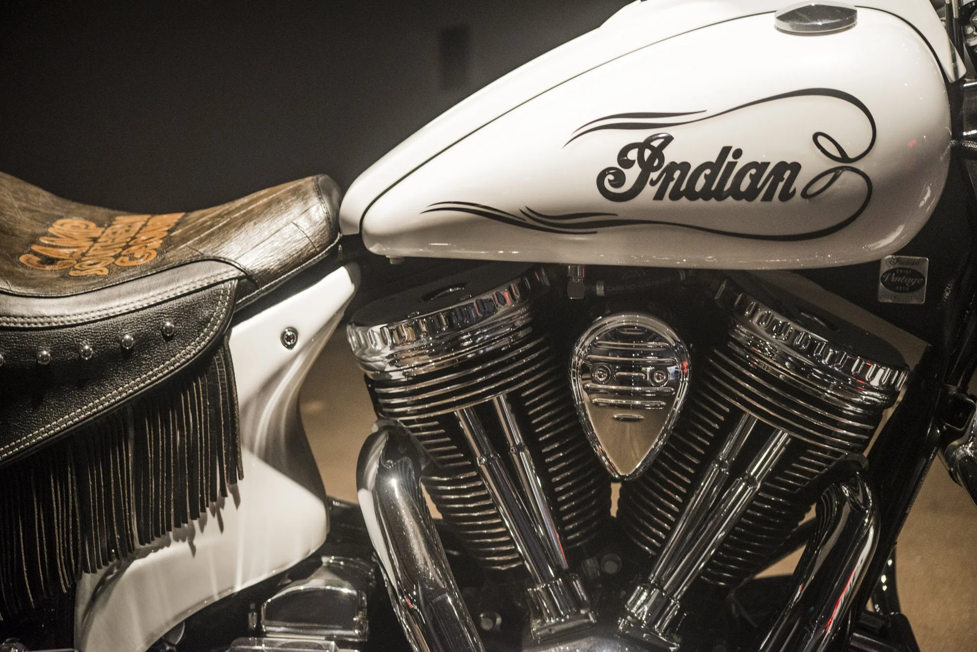Close-up of a white Indian motorcycle with black and chrome engine components and a tan leather seat with fringe details.