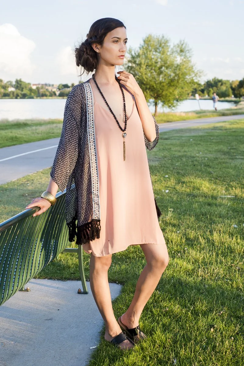 A woman standing outdoors near a lake, wearing a light pink dress, black sandals, and a black beaded necklace, with her hand on a park bench.