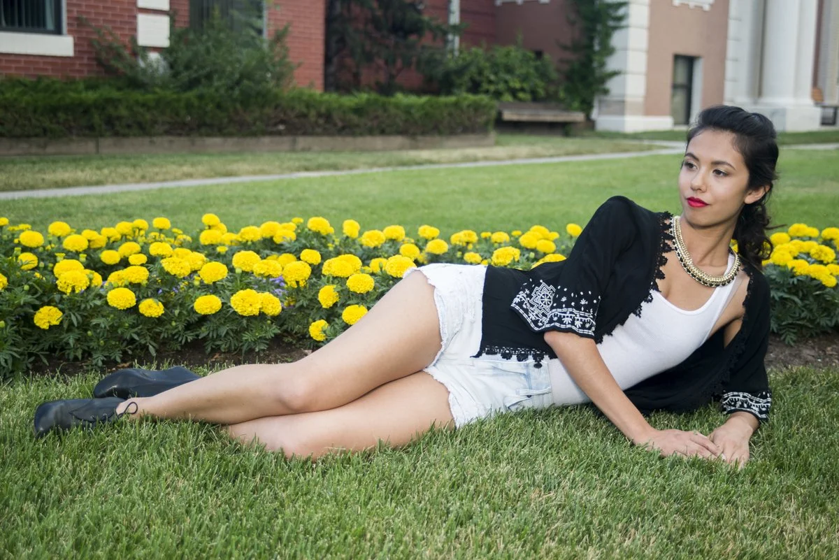 A woman lying on the grass beside a flower bed of yellow marigolds in a park with a red brick building in the background.