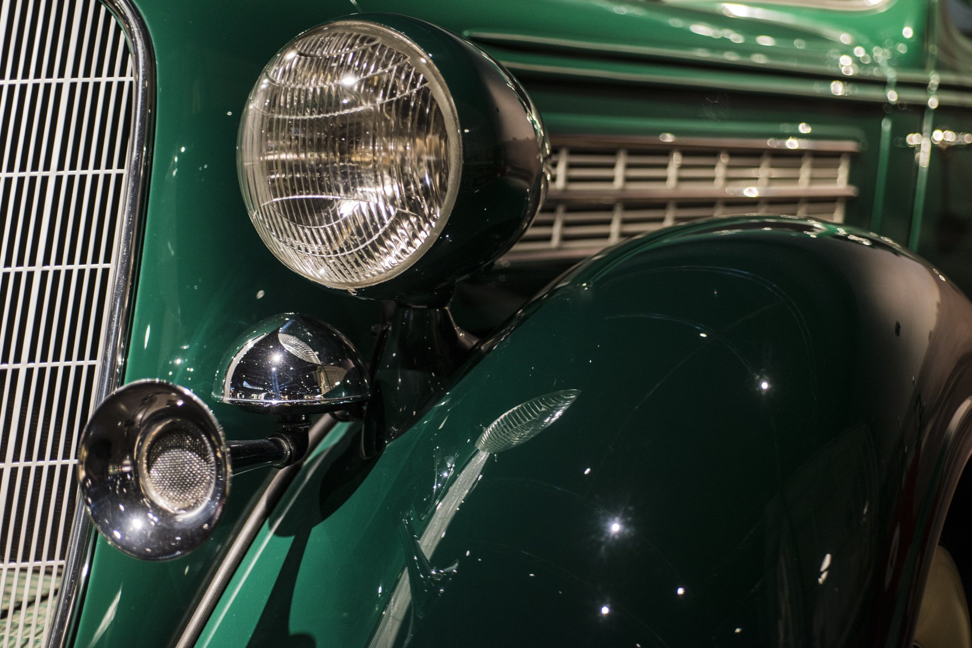 Close-up of a vintage green car's front end, featuring a large round headlight, a smaller auxiliary light, and a vertical chrome grille.