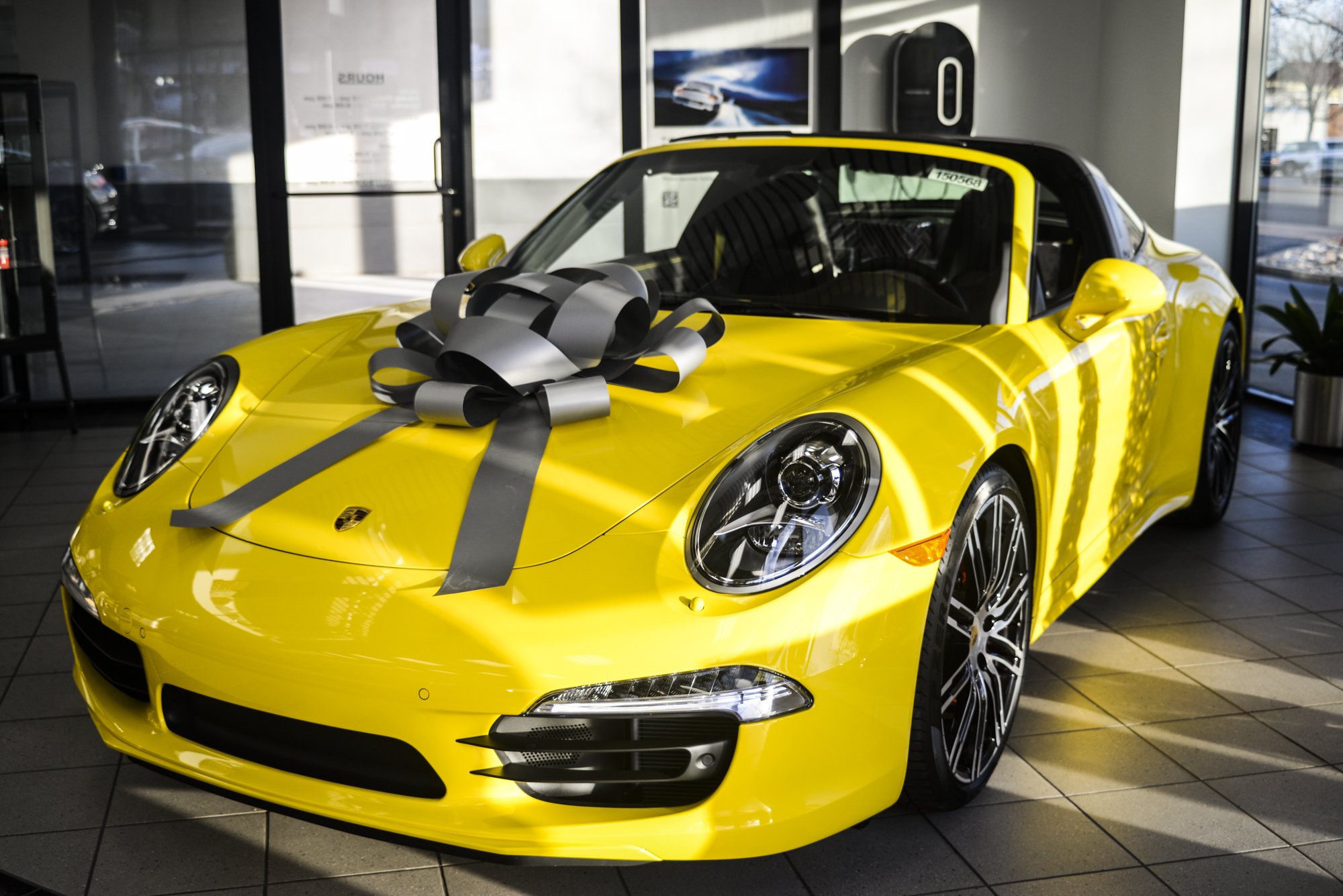 A bright yellow sports car with a large gray gift bow on the hood, displayed inside a car dealership.