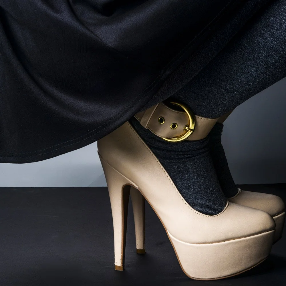 Close-up of beige high-heeled platform shoes with black socks and a beige belt with a large gold buckle, worn by a person dressed in dark clothing.